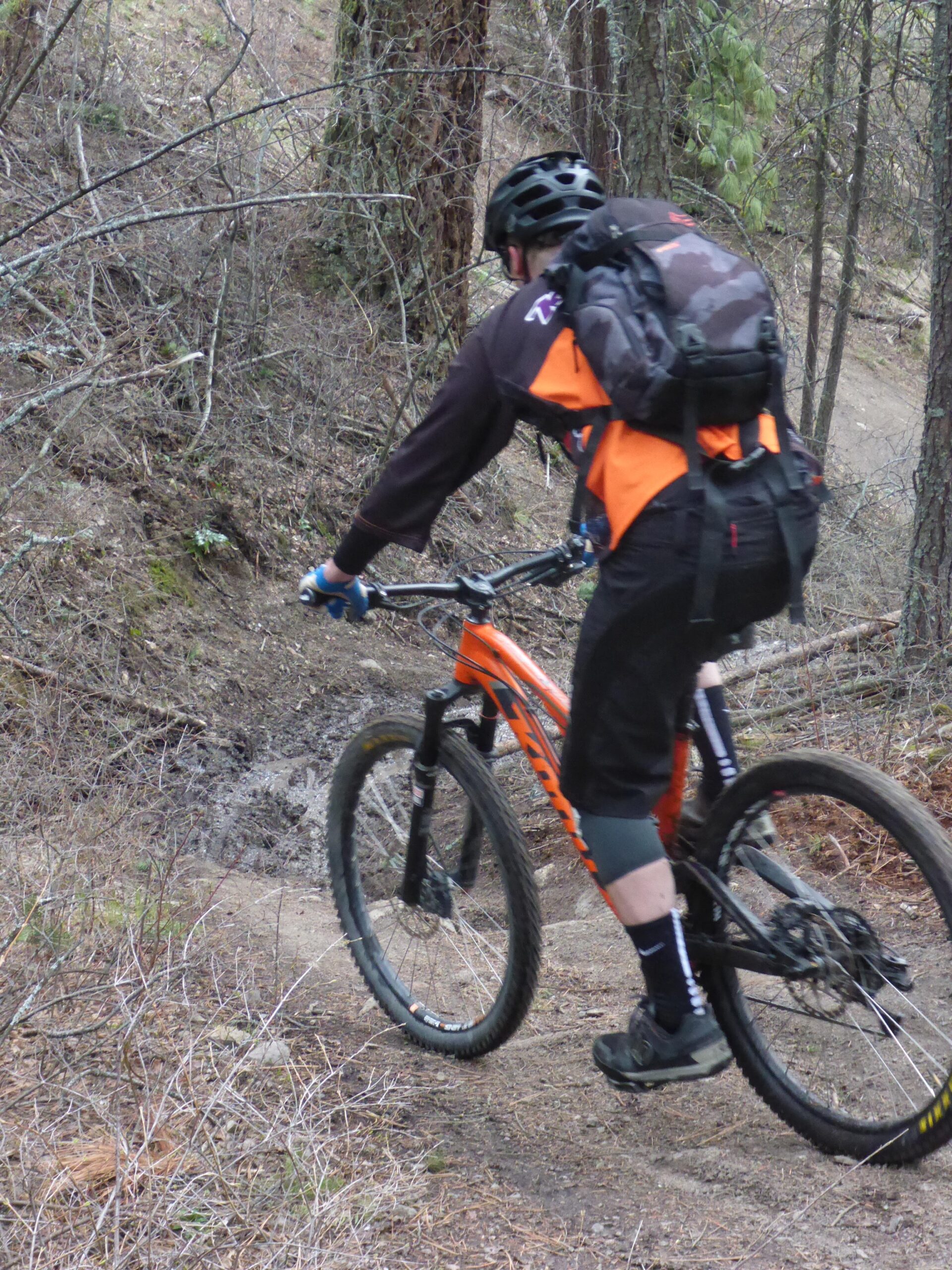 A mountain biker wearing a black and orange jersey and a helmet navigates a narrow, rocky trail surrounded by trees and underbrush. The bike is predominantly orange, and the rider is focused on the path ahead, showcasing an outdoor recreational activity in a natural setting. Smith Creek mountain bike trail.