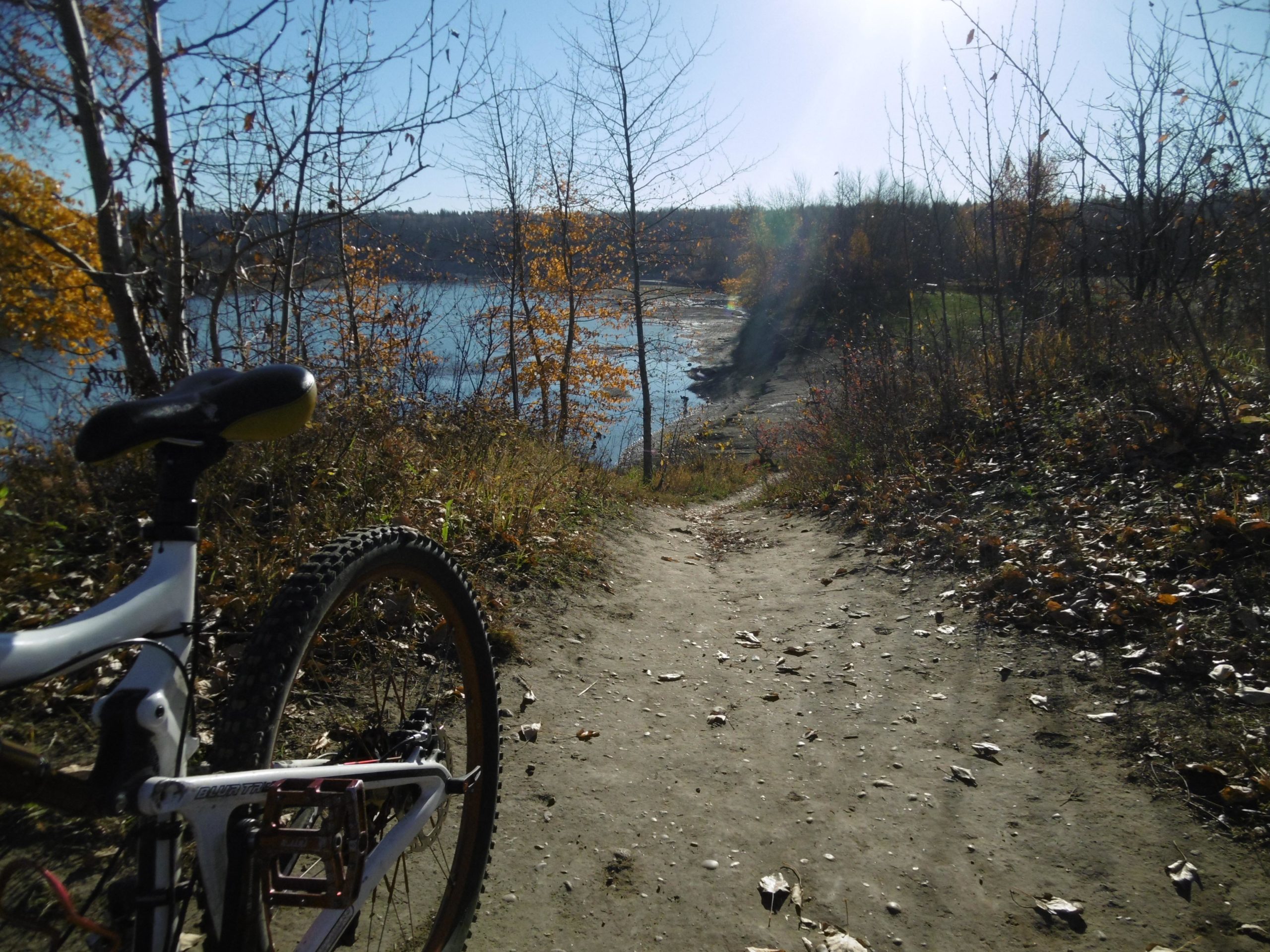 A mountain bike is positioned on a dirt path overlooking a tranquil body of water surrounded by trees. The scene is set in autumn with colorful leaves on the trees, and the sunlight creates a bright and warm atmosphere. Terwillegar Park mountain bike trail.