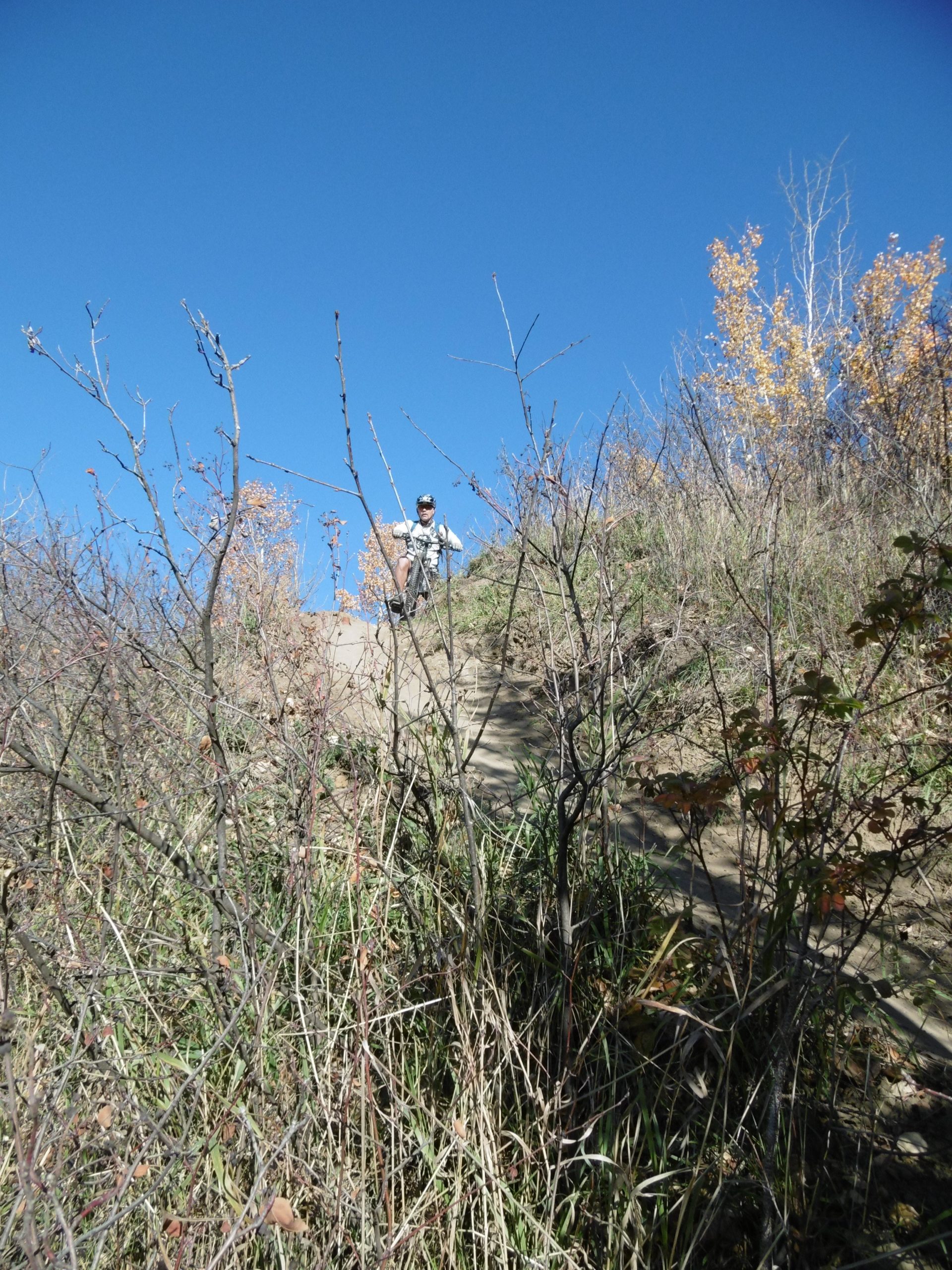 A person sitting on a hillside surrounded by sparse vegetation, with a clear blue sky above. The scene captures a tranquil outdoor setting in nature. Terwillegar Park mountain bike trail.