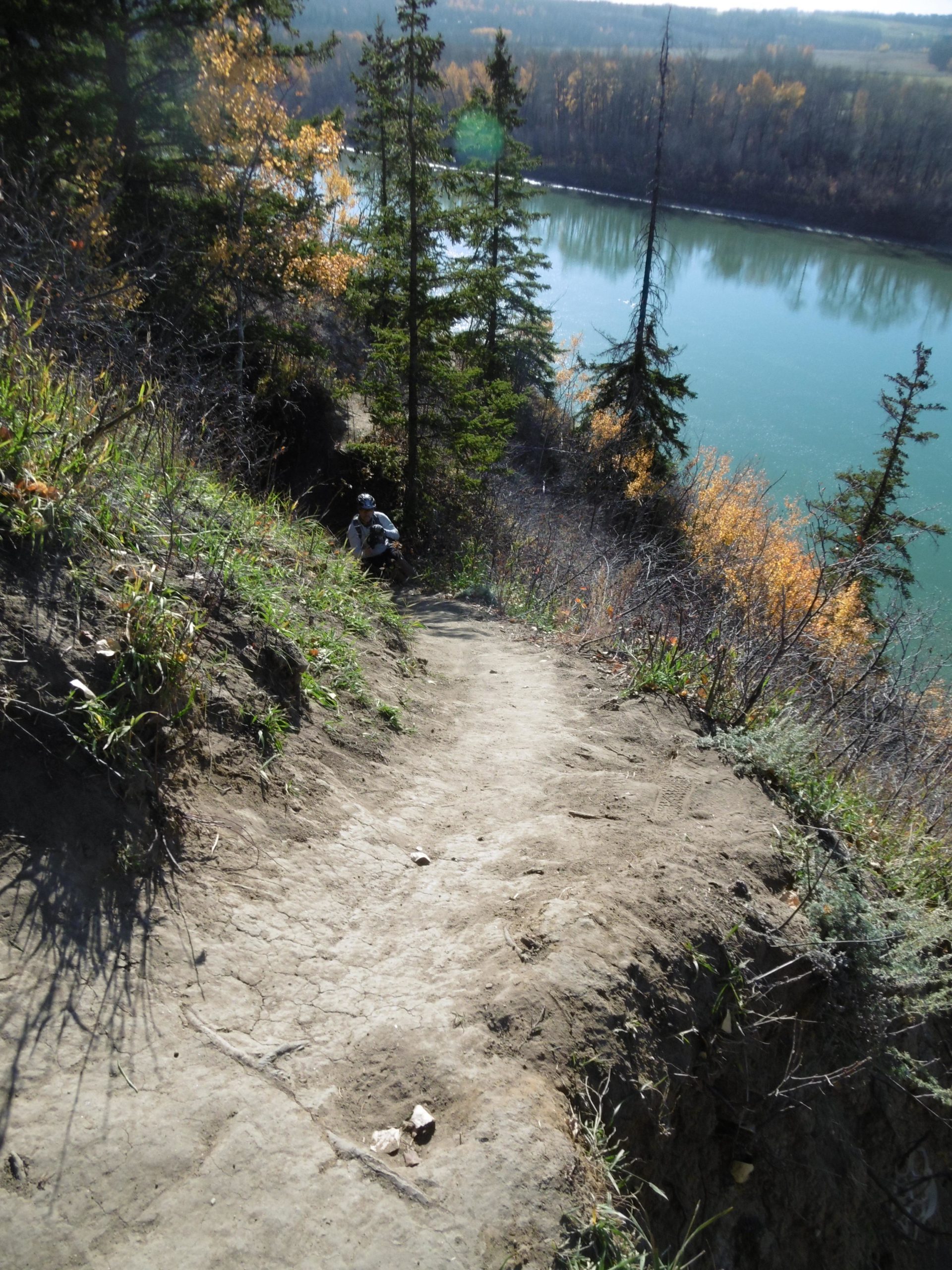 A winding dirt path leads down a hillside, surrounded by greenery and trees with autumn foliage. In the foreground, the path is visible, slightly worn and crisscrossed, while in the background, a river reflects the blue sky. The scene conveys a sense of nature and outdoor adventure. Terwillegar Park mountain bike trail.