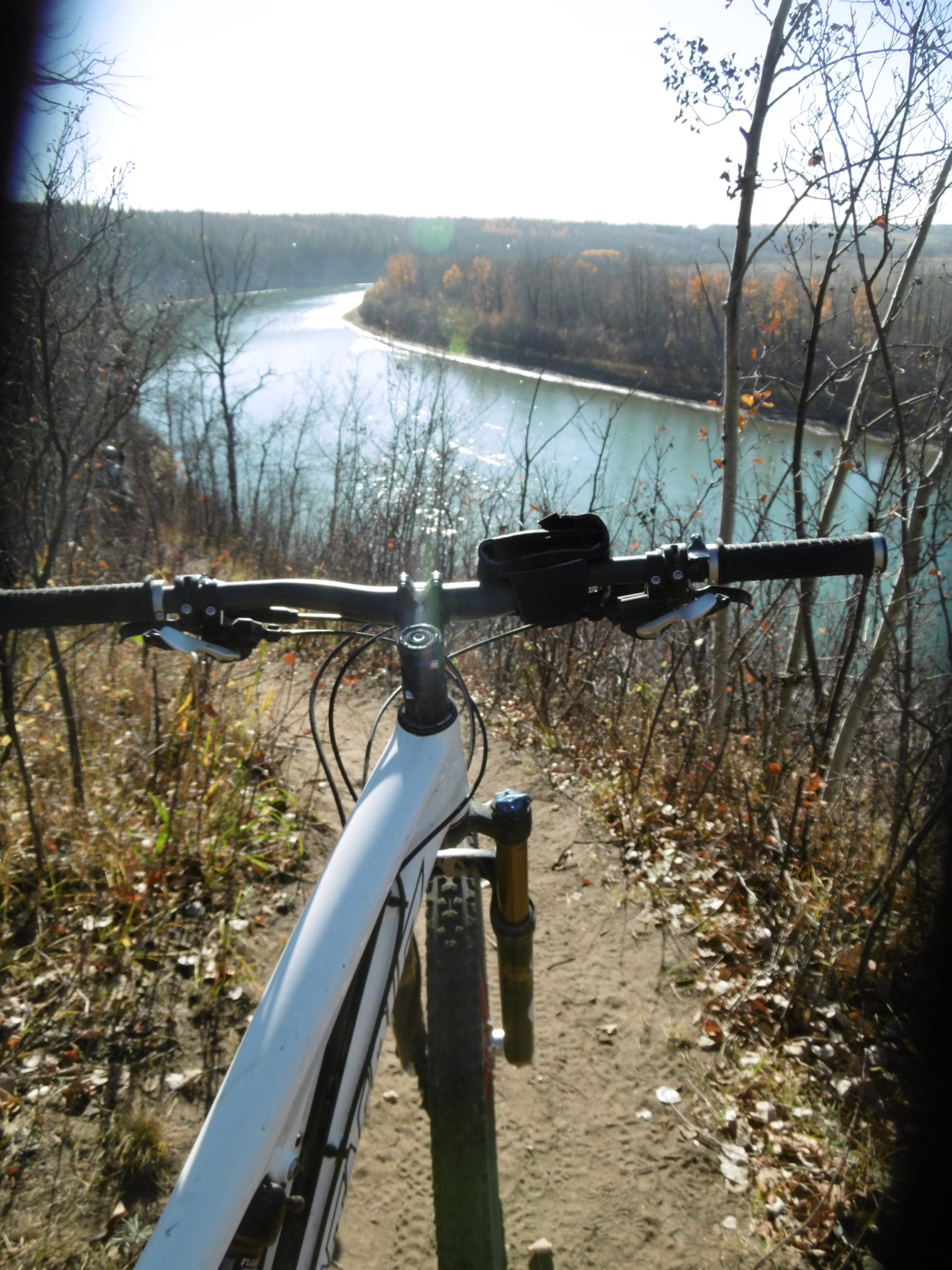 A view from the handlebars of a mountain bike, overlooking a winding river surrounded by trees with autumn foliage. The sun is shining brightly in a clear blue sky, illuminating the dirt path and nearby shrubs. Terwillegar Park mountain bike trail.