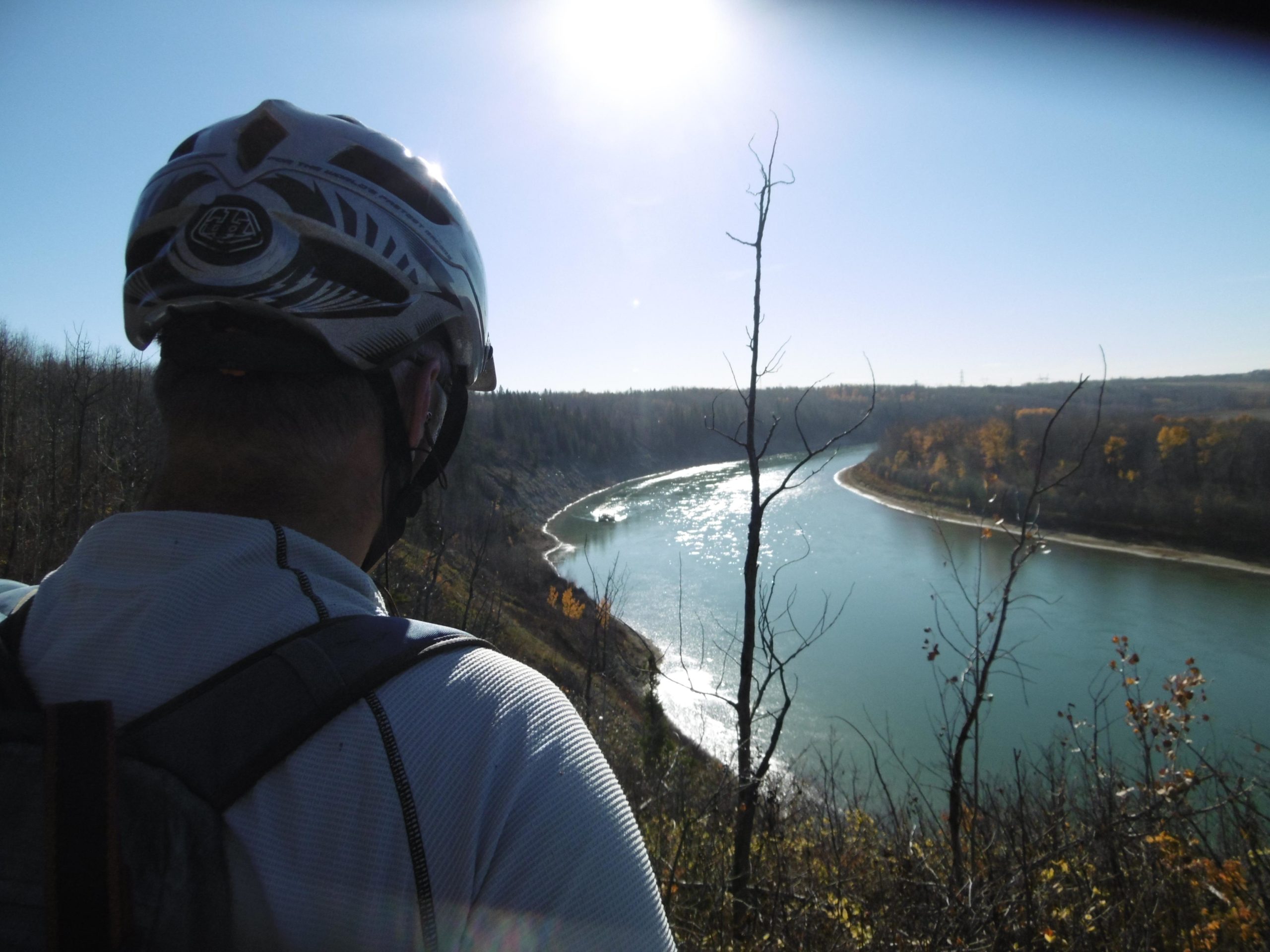 A person wearing a cycling helmet overlooks a river winding through a landscape of trees and hills. The sunlight reflects off the water, and it is a clear blue sky. Autumn foliage is visible in the background. Terwillegar Park mountain bike trail.