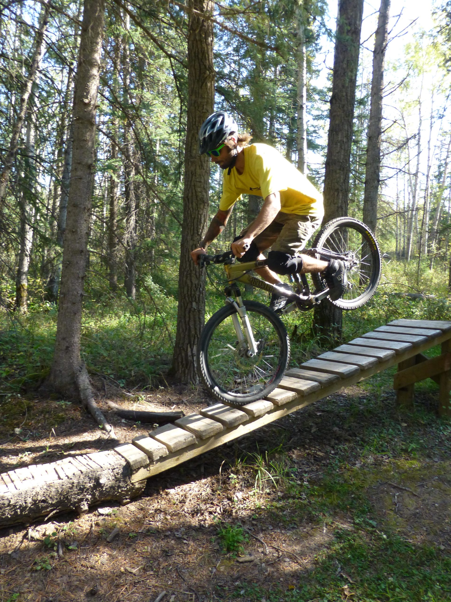 A person wearing a yellow shirt and helmet rides a mountain bike over a narrow wooden trail bridge in a forested area, with trees and greenery surrounding them. Hinton Skills Park mountain bike trail.