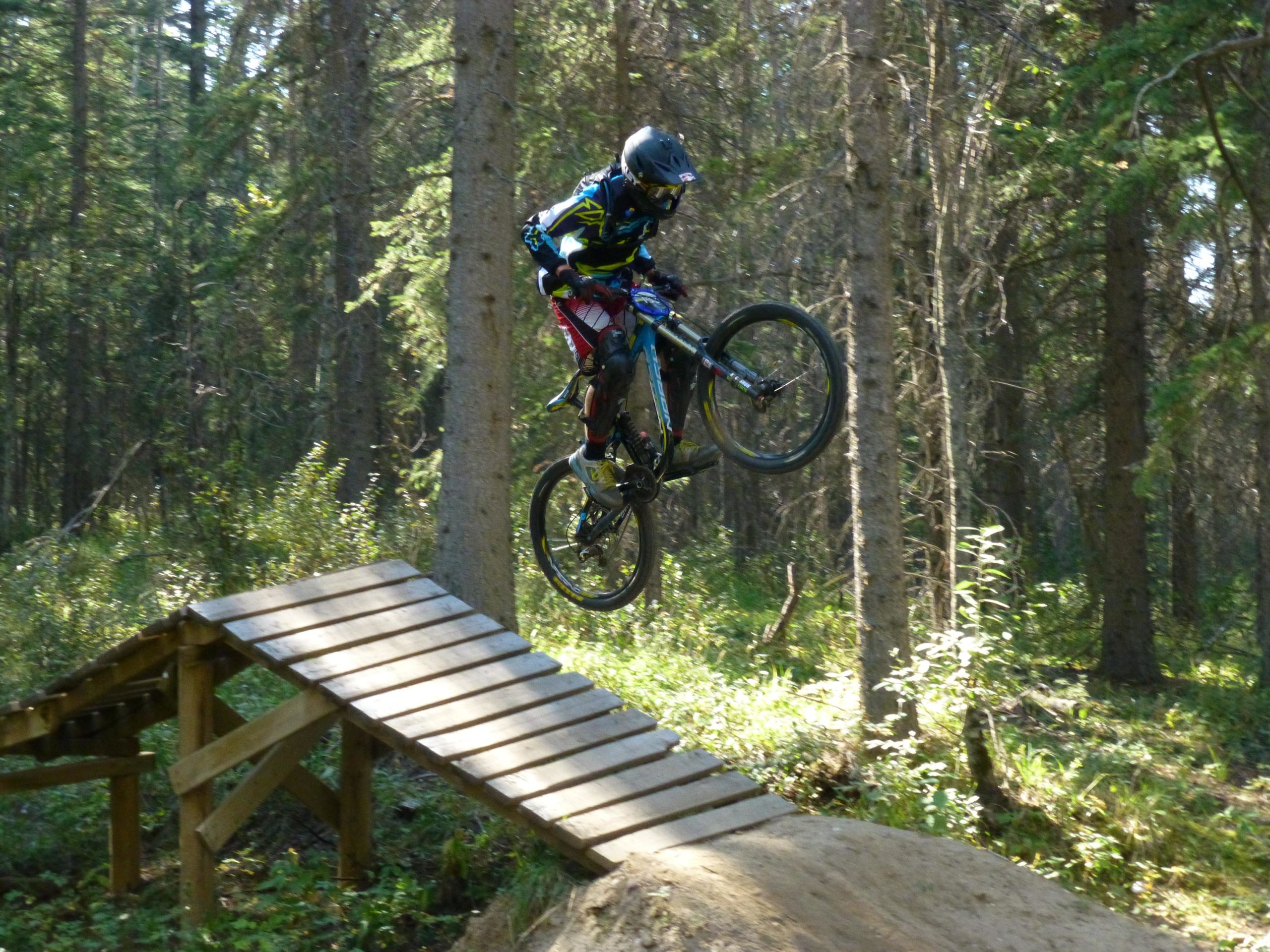 A mountain biker wearing a helmet and protective gear jumps off a wooden ramp in a forested area, surrounded by trees and greenery. The bike is airborne, showcasing the rider's skill in off-road cycling. Hinton Skills Park mountain bike trail.
