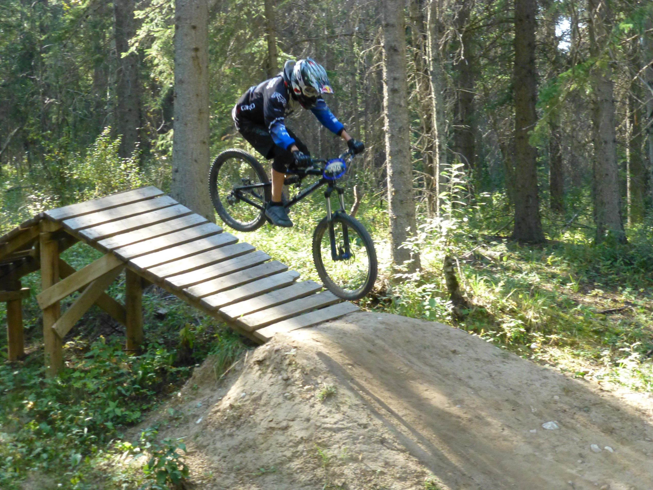 A cyclist wearing protective gear jumps off a wooden ramp in a forested area, with trees and greenery surrounding the scene. Hinton Skills Park mountain bike trail.