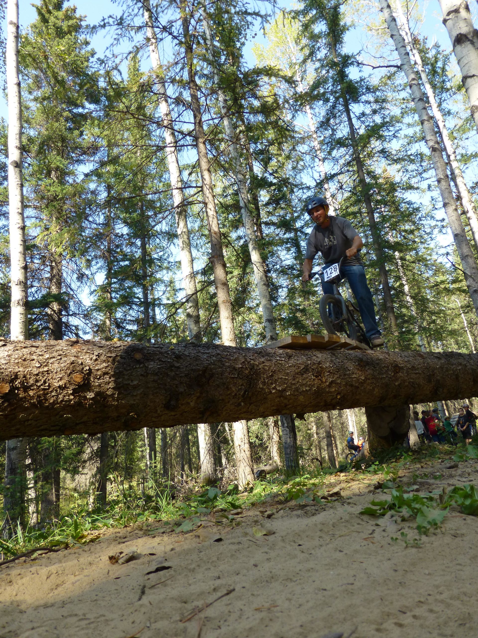 A mountain biker performs a stunt on a log in a forest, with tall trees surrounding him. The rider is wearing a helmet and a grey shirt, concentrating on maintaining balance as he rides along the log. In the background, some spectators can be seen watching the action. Hinton Skills Park mountain bike trail.