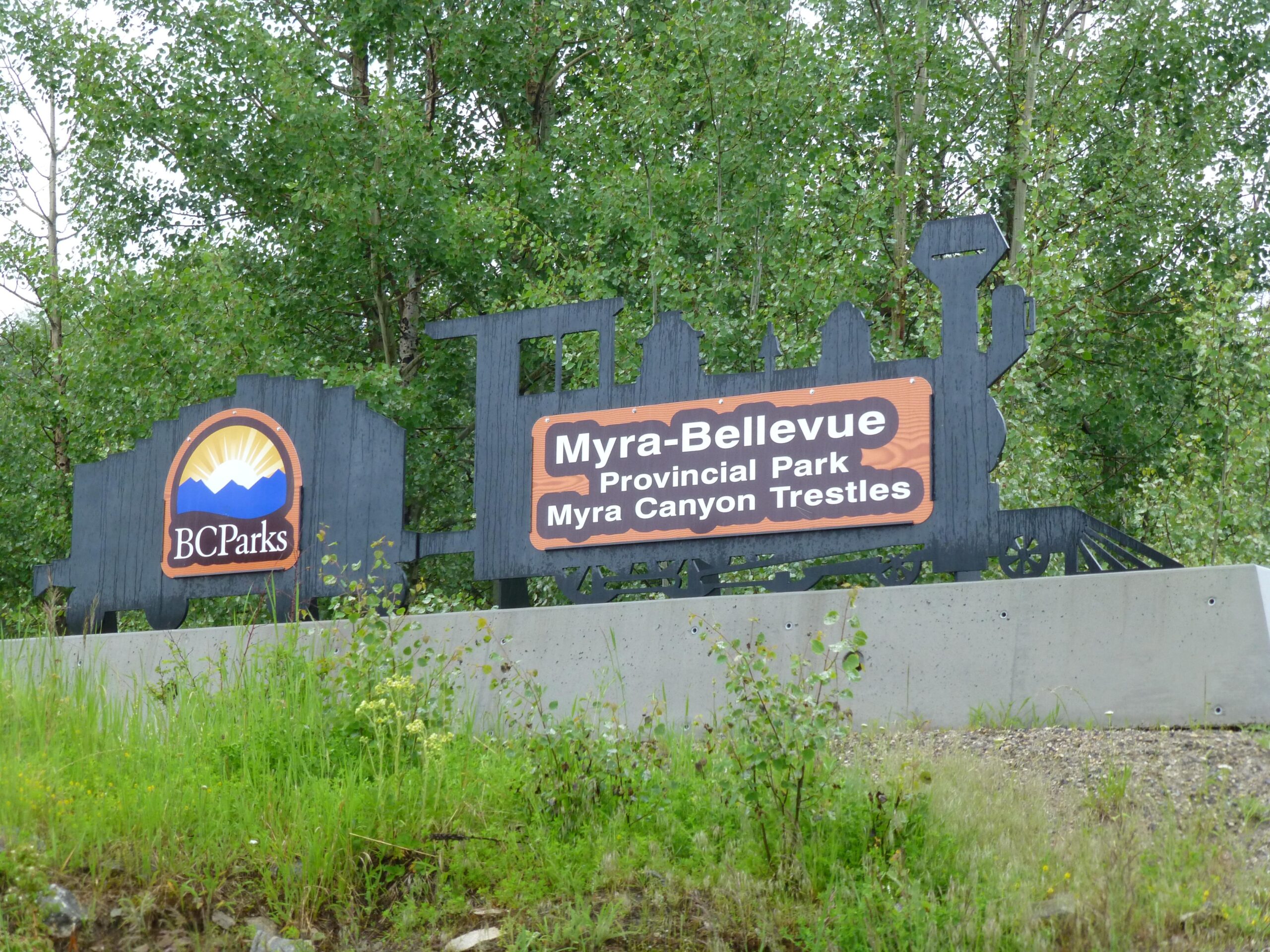 A wooden sign for Myra-Bellevue Provincial Park, featuring the BC Parks logo. The sign includes the park's name and mentions "Myra Canyon Trestles," with a background of green foliage and trees. Kettle Valley Railway Trail (KVR) Myra Canyon to Penticton return mountain bike trail.