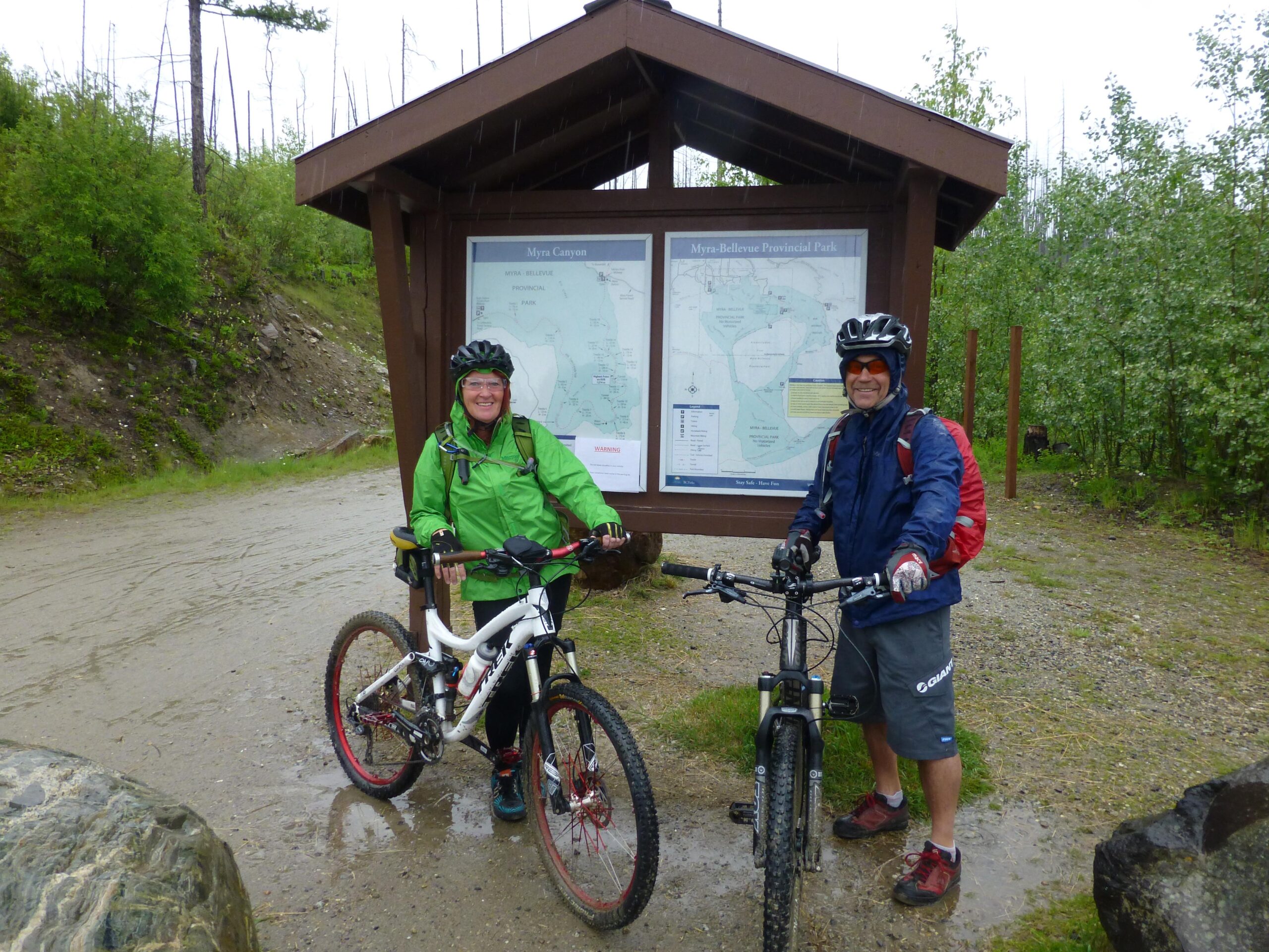 Two mountain bikers stand beside a map display at the entrance of Myra-Bellevue Provincial Park. One rider, wearing a green rain jacket and helmet, leans on a white mountain bike, while the other, dressed in a blue jacket with a red backpack, smiles next to a black mountain bike. The ground is wet, indicating recent rain, and the surrounding area features greenery and a few trees. Kettle Valley Railway Trail (KVR) Myra Canyon to Penticton return mountain bike trail.