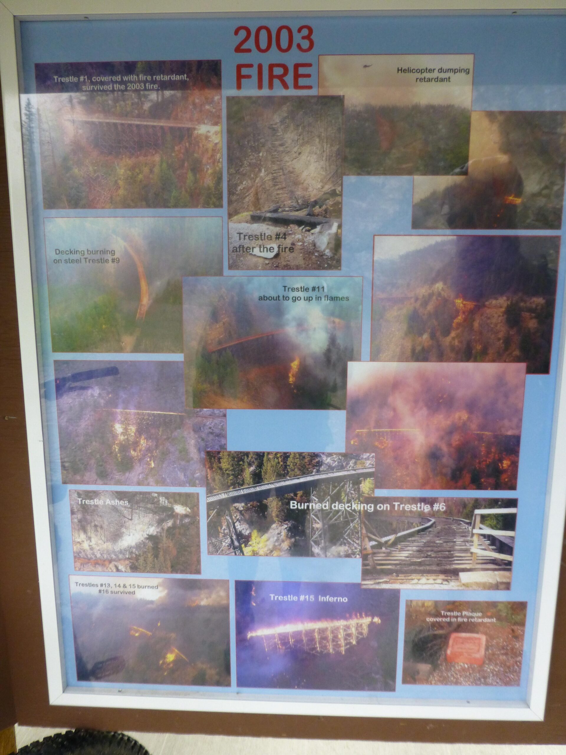 A collage of photos documenting the impact of a wildfire in 2003, featuring images of damaged and surviving trestles, alongside descriptions of each. The photos illustrate scenes of burned decking, trestles engulfed in flames, and firefighting efforts, including a helicopter dropping fire retardant. The display highlights the resilience of some structures, marked with labels such as "Trestle #1 survived" and "Trestles #13, 14 & 15 burned," providing a visual history of the fire's effects on the landscape and infrastructure. Kettle Valley Railway Trail (KVR) Myra Canyon to Penticton return mountain bike trail.