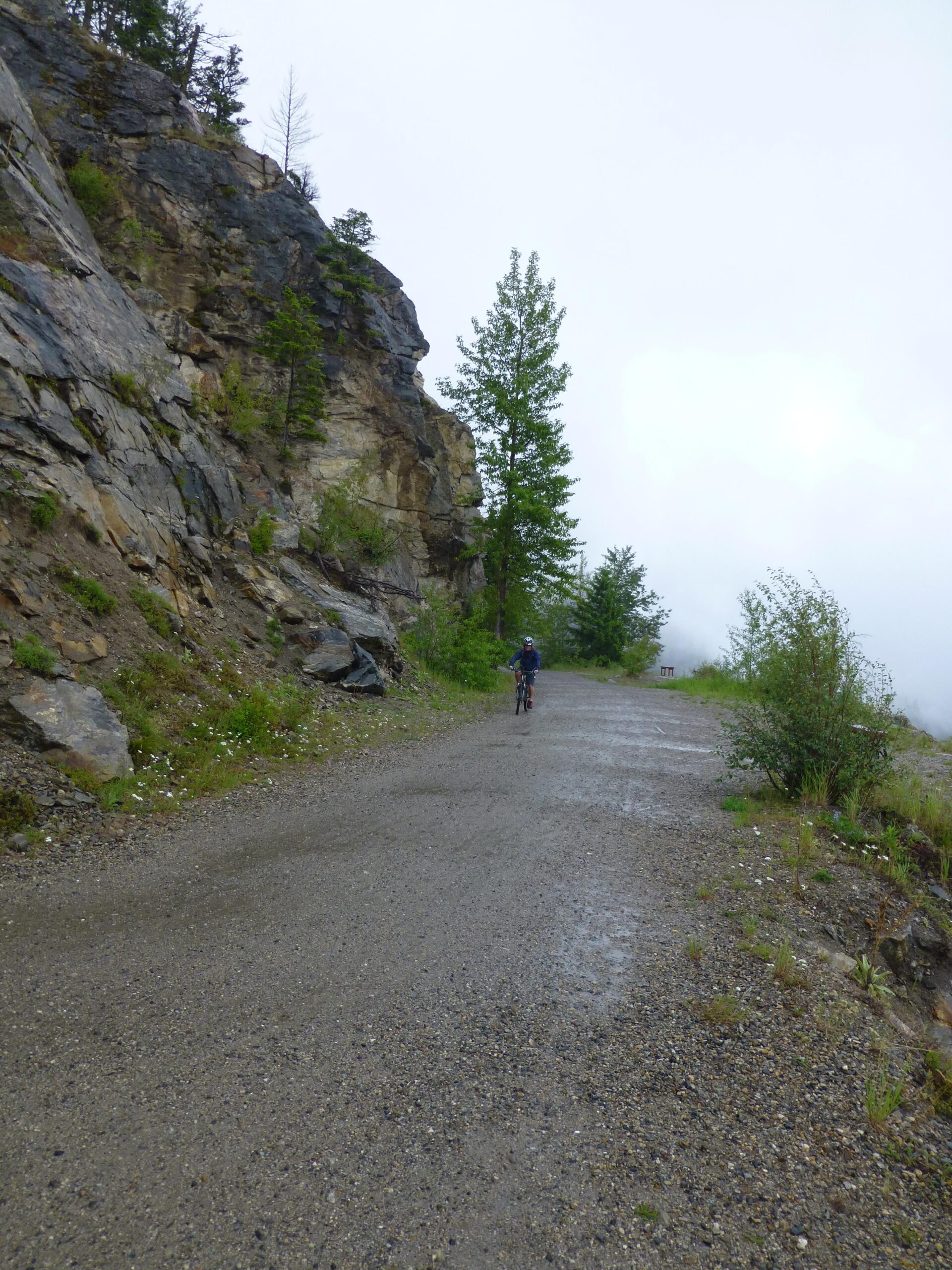A cyclist riding on a gravel path alongside a rocky cliff, surrounded by green trees and misty weather, with cloudy skies above. Kettle Valley Railway Trail (KVR) Myra Canyon to Penticton return mountain bike trail.