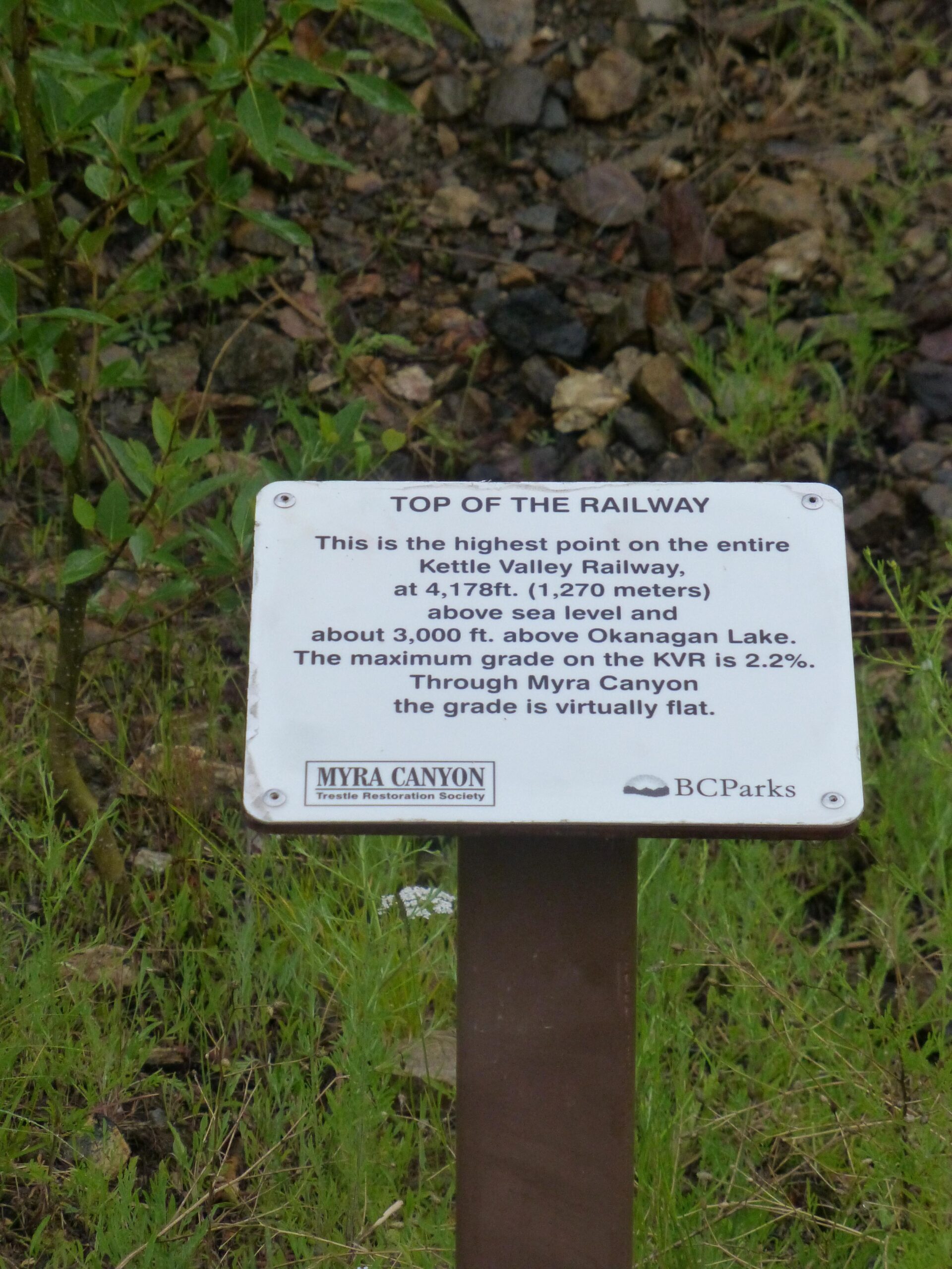 Information sign at the top of the Kettle Valley Railway, stating it is the highest point at 4,178 feet (1,270 meters) above sea level, near Okanagan Lake. The sign includes details about the maximum grade of the railway and its location in Myra Canyon. Surrounded by greenery and rocky terrain. Kettle Valley Railway Trail (KVR) Myra Canyon to Penticton return mountain bike trail.