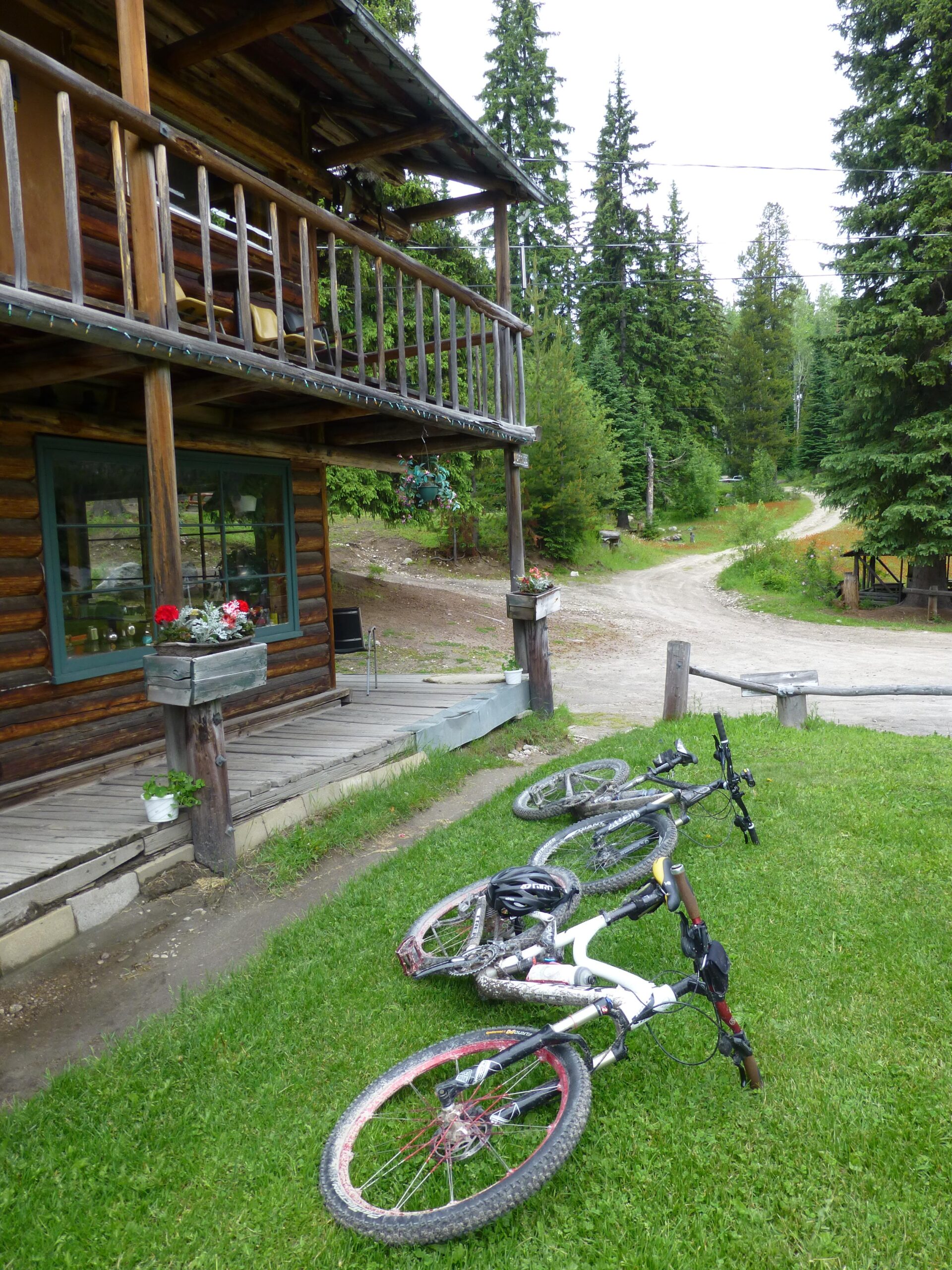 A rustic log cabin surrounded by tall evergreen trees, with a dirt road leading into the forest. In the foreground, four mountain bikes are resting on the lush green grass, showing signs of mud and use. A flower box and potted plants decorate the cabin’s porch, adding a touch of color to the natural setting. Kettle Valley Railway Trail (KVR) Myra Canyon to Penticton return mountain bike trail.