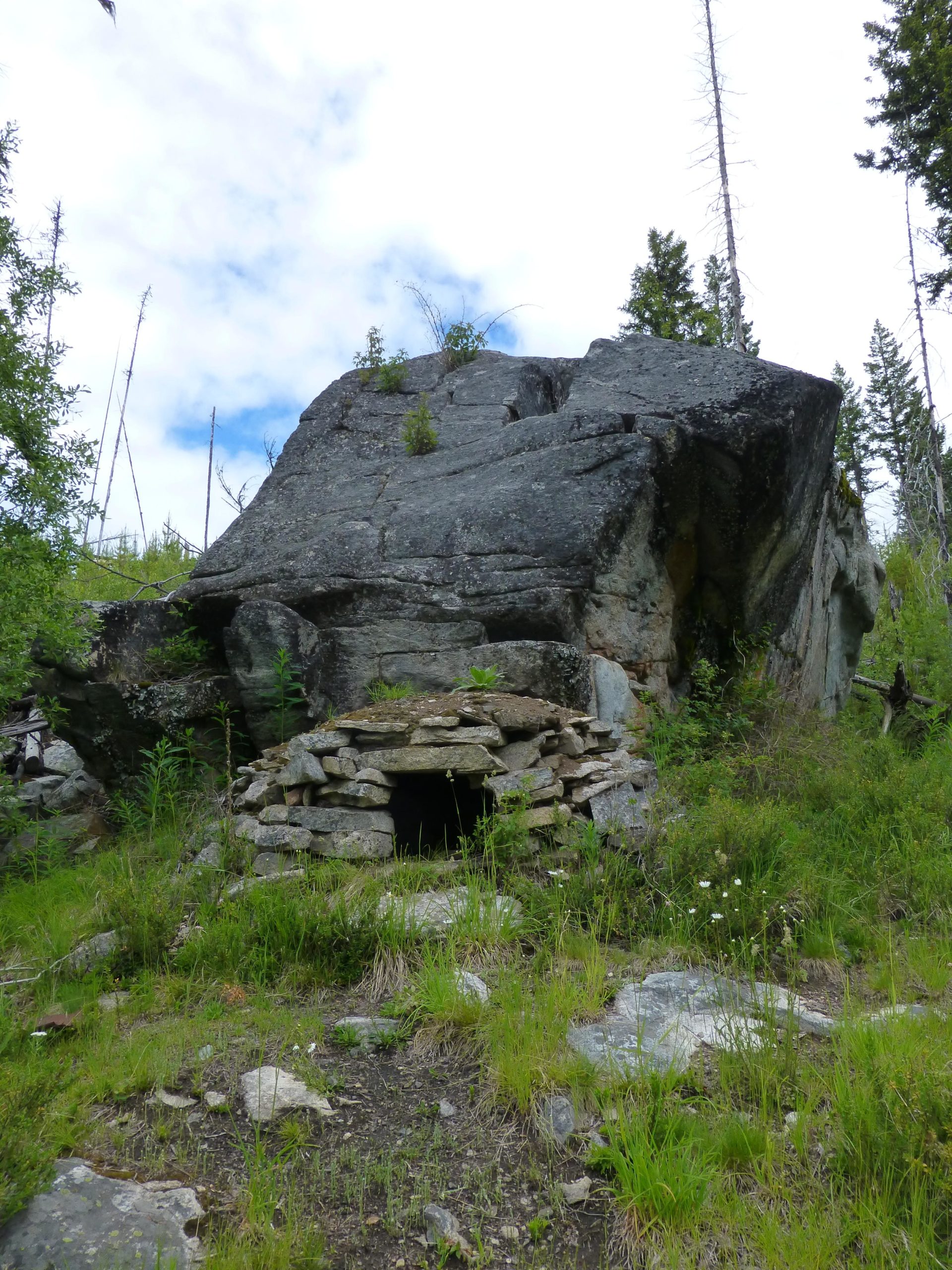 A small stone structure built into the base of a large boulder, surrounded by green grass and small plants. The entrance of the structure is dark and partially obscured. Pine trees and a cloudy sky are visible in the background. Kettle Valley Railway Trail (KVR) Myra Canyon to Penticton return mountain bike trail.