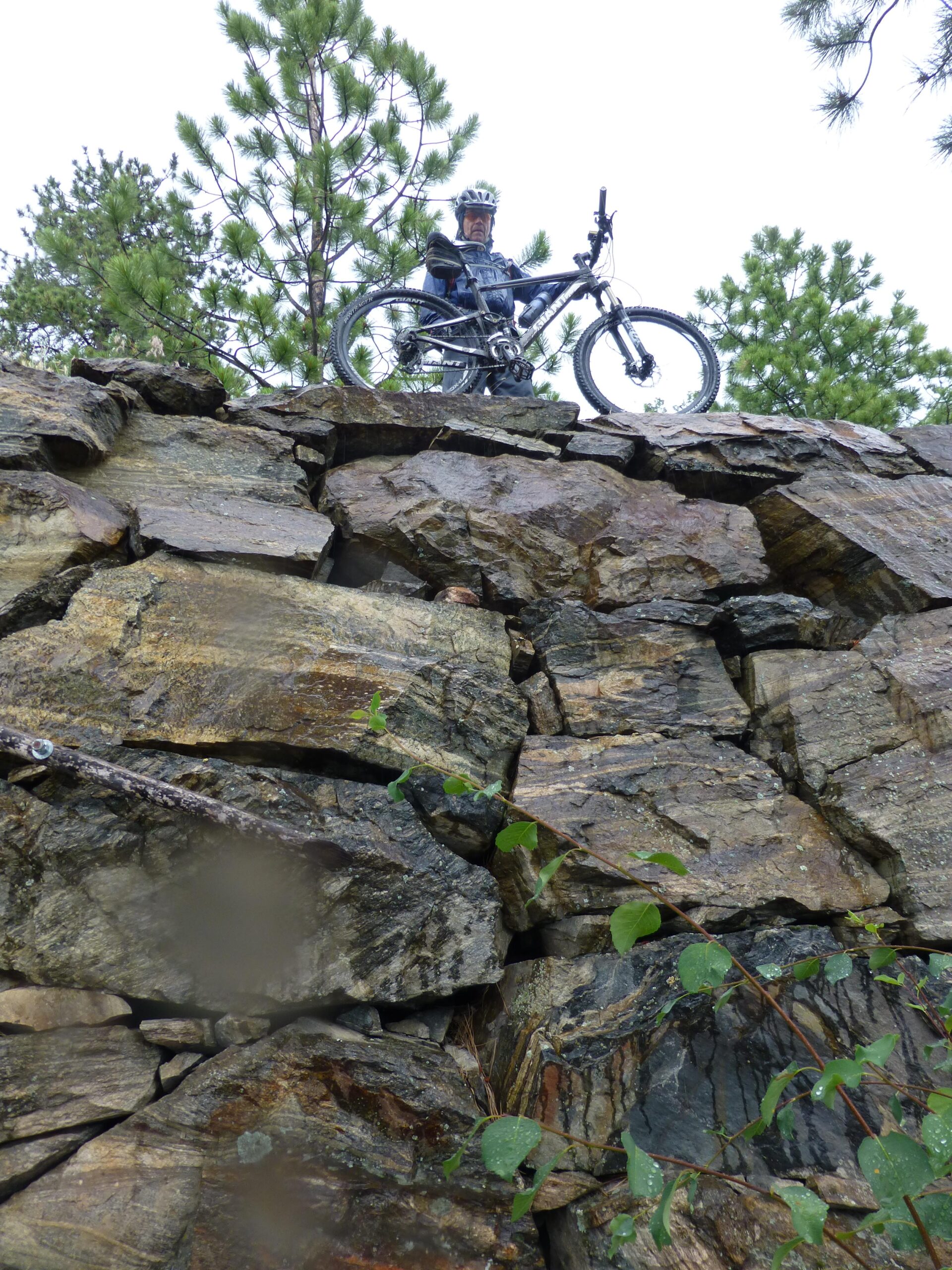 A mountain biker standing atop a rocky outcrop with a forest of pine trees in the background, holding a mountain bike. The scene depicts an adventurous outdoor activity in a natural setting, with rain-soaked rocks and greenery. Kettle Valley Railway Trail (KVR) Myra Canyon to Penticton return mountain bike trail.