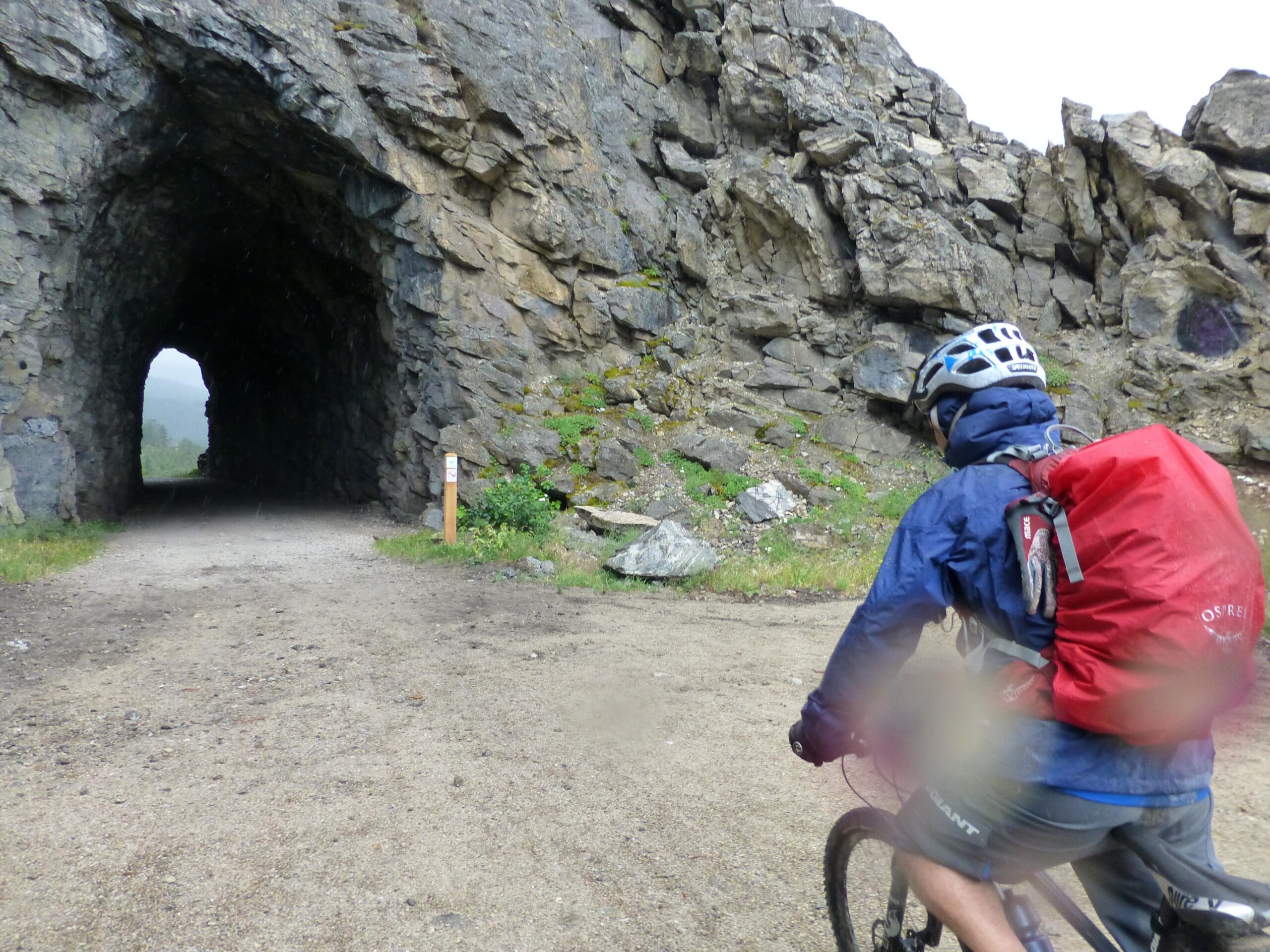A cyclist wearing a helmet and a blue rain jacket rides on a dirt path leading toward a rocky tunnel. The tunnel, formed by large boulders, opens up to a lush green landscape in the background, indicating an outdoor adventure in a mountainous area. Light rain is falling, adding a misty atmosphere to the scene. Kettle Valley Railway Trail (KVR) Myra Canyon to Penticton return mountain bike trail.