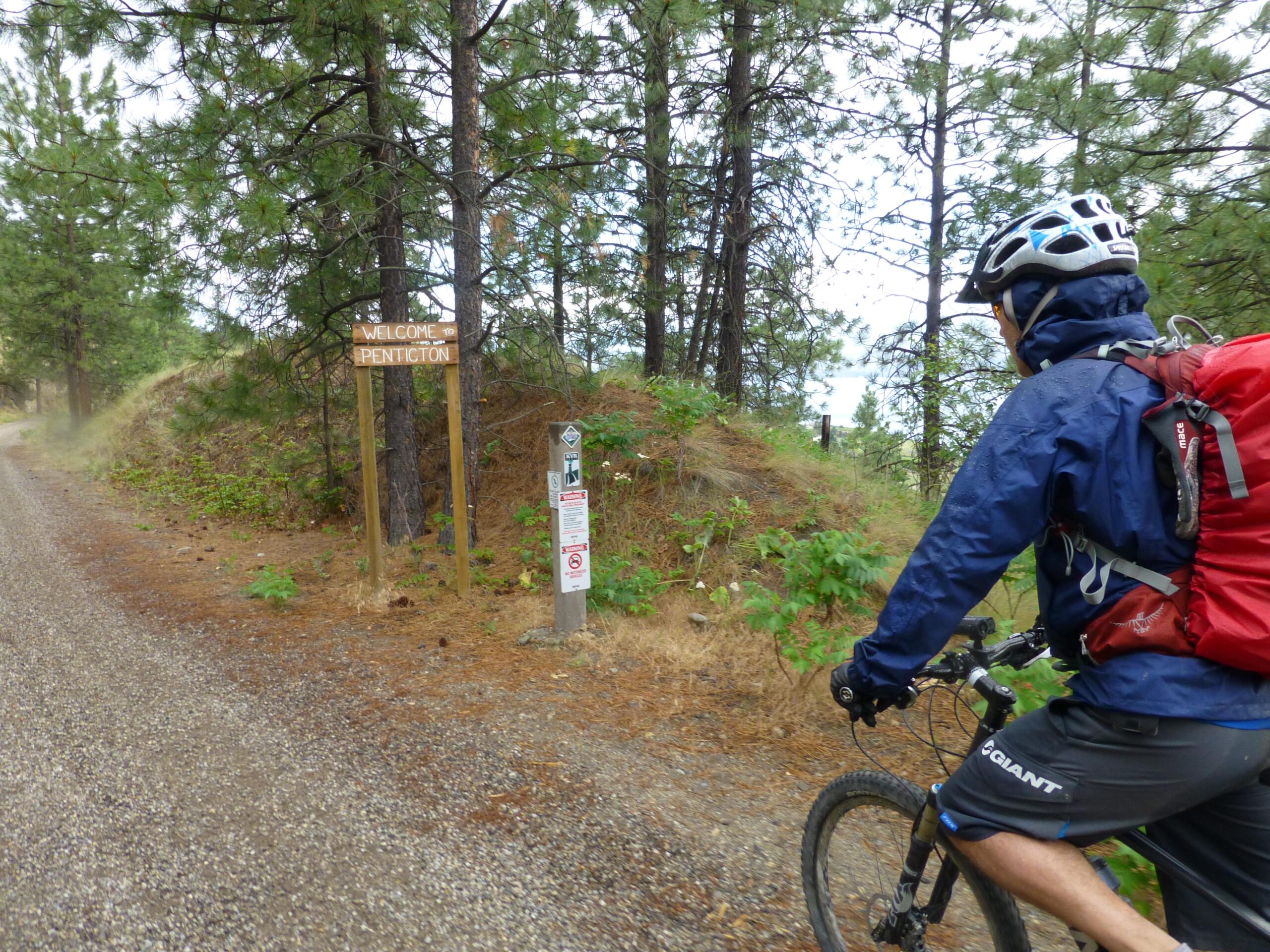 A cyclist wearing a helmet and waterproof jacket rides along a gravel path in a forested area, with a "Welcome to Penticton" sign visible on the side. Signs indicating trail regulations are also present. The environment appears lush and green, typical of a pleasant outdoor location. Kettle Valley Railway Trail (KVR) Myra Canyon to Penticton return mountain bike trail.