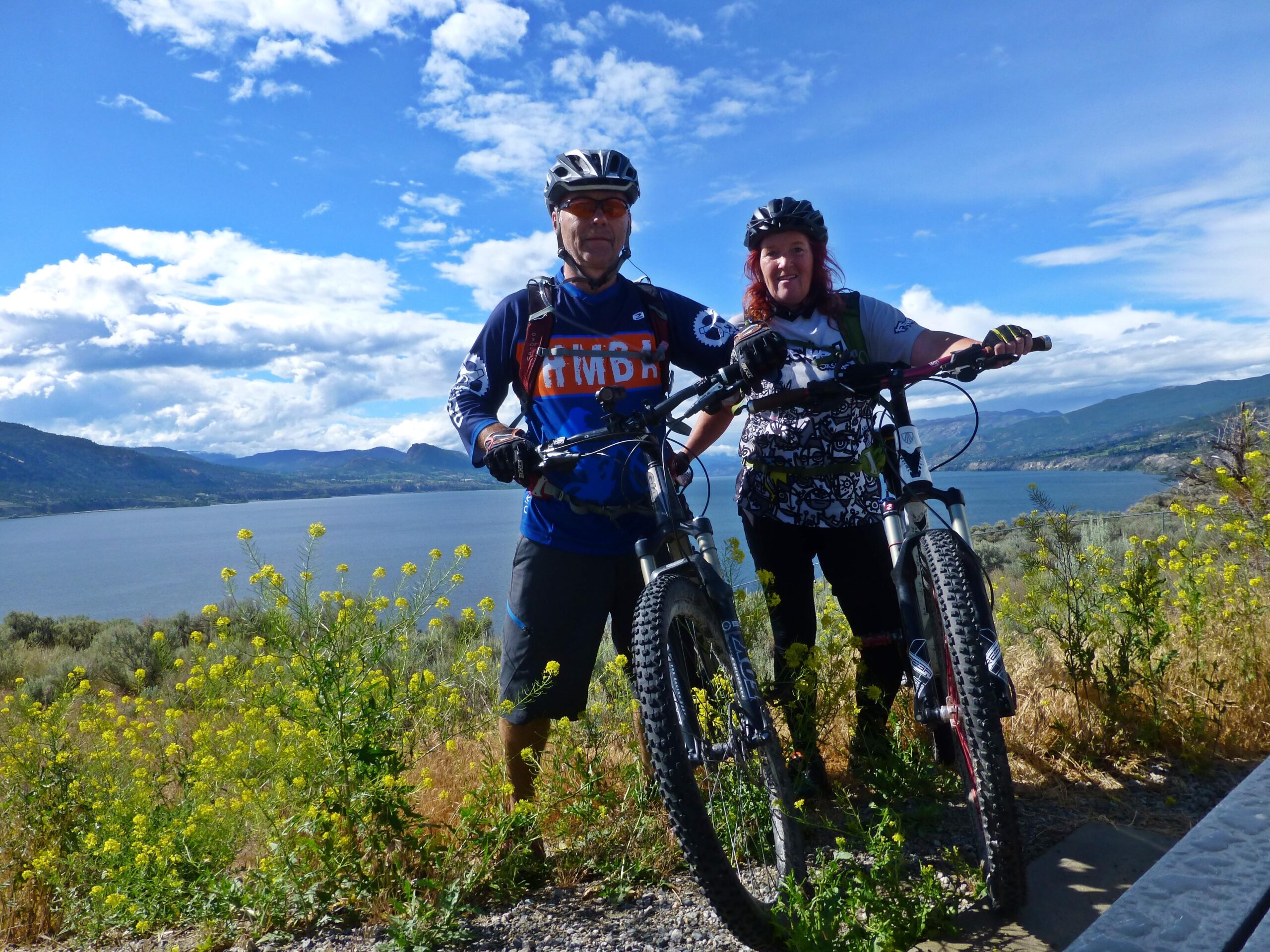 Two mountain bikers stand beside their bikes on a scenic overlook. They are smiling and dressed in cycling gear, with a backdrop of a lake and rolling hills under a partly cloudy sky. Bright yellow wildflowers and green foliage frame the foreground. Kettle Valley Railway Trail (KVR) Myra Canyon to Penticton return mountain bike trail.