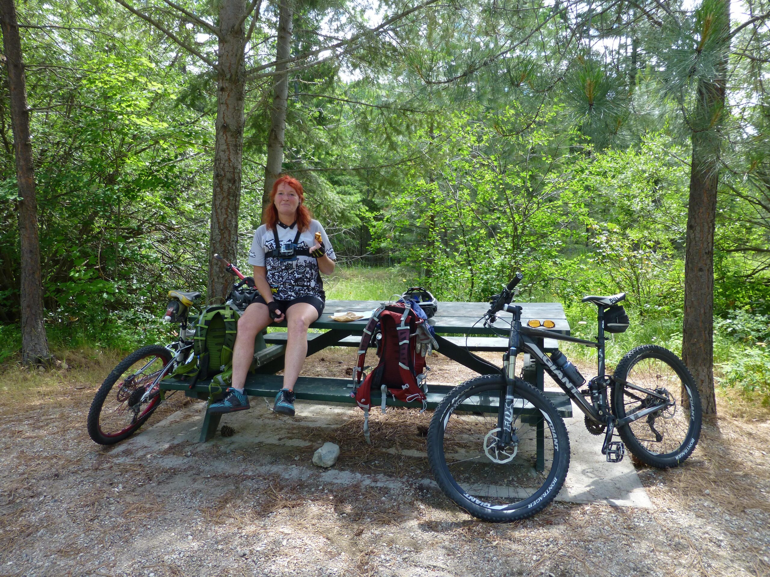 A person with red hair sitting at a picnic table in a wooded area, holding food and smiling. Two bicycles are parked nearby, and the surrounding environment features lush greenery. Kettle Valley Railway Trail (KVR) Myra Canyon to Penticton return mountain bike trail.
