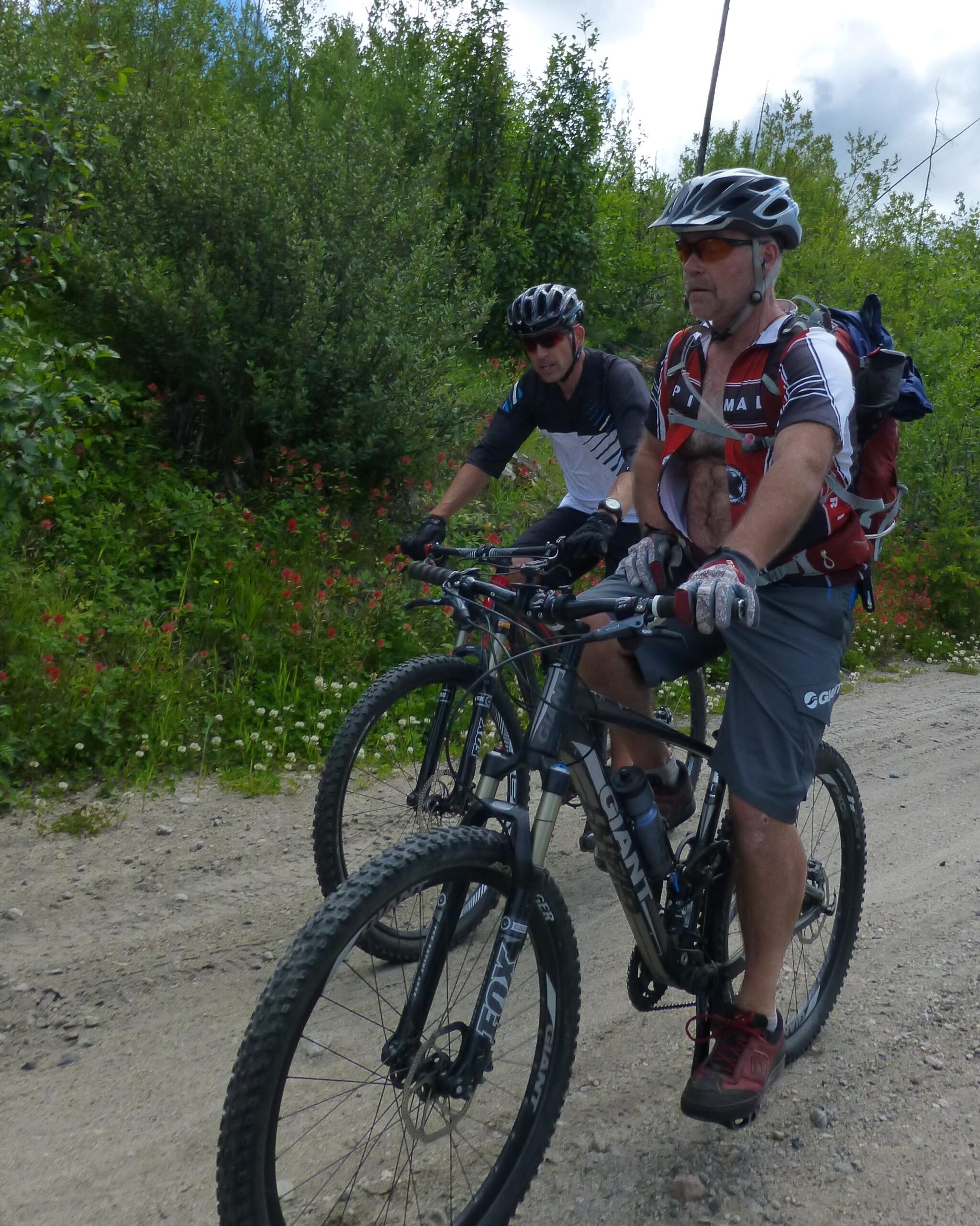 Two mountain bikers are riding on a gravel path surrounded by greenery and wildflowers. One biker is in the foreground, wearing a helmet and sunglasses, with a backpack and gloves, while the second biker is slightly behind, dressed in a lighter top. The scene is bright and captures the essence of outdoor adventure. Kettle Valley Railway Trail (KVR) Myra Canyon to Penticton return mountain bike trail.