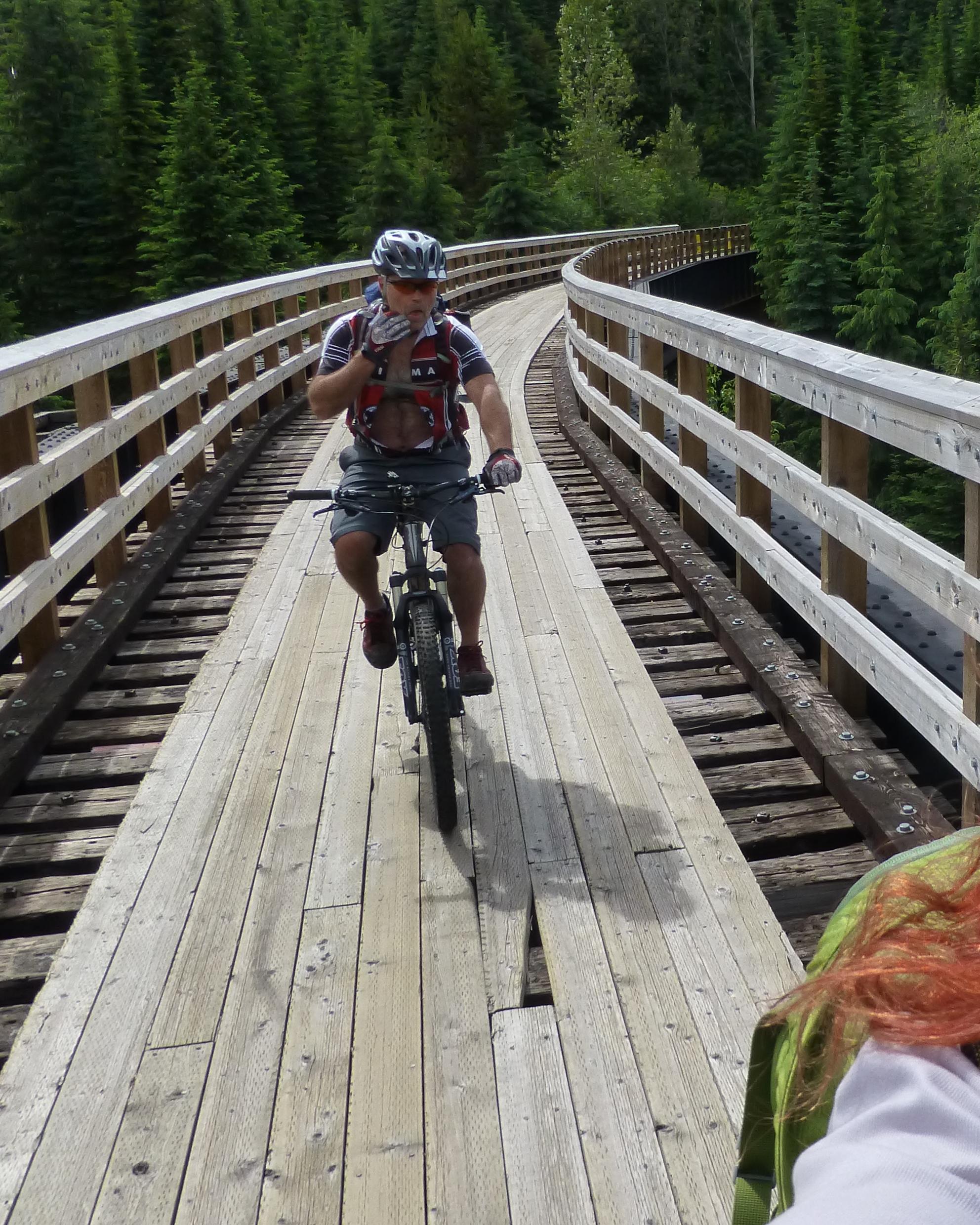A cyclist on a wooden bridge surrounded by lush green trees, navigating a curved path. The rider is wearing a helmet and a backpack, appearing to pause while riding. Kettle Valley Railway Trail (KVR) Myra Canyon to Penticton return mountain bike trail.
