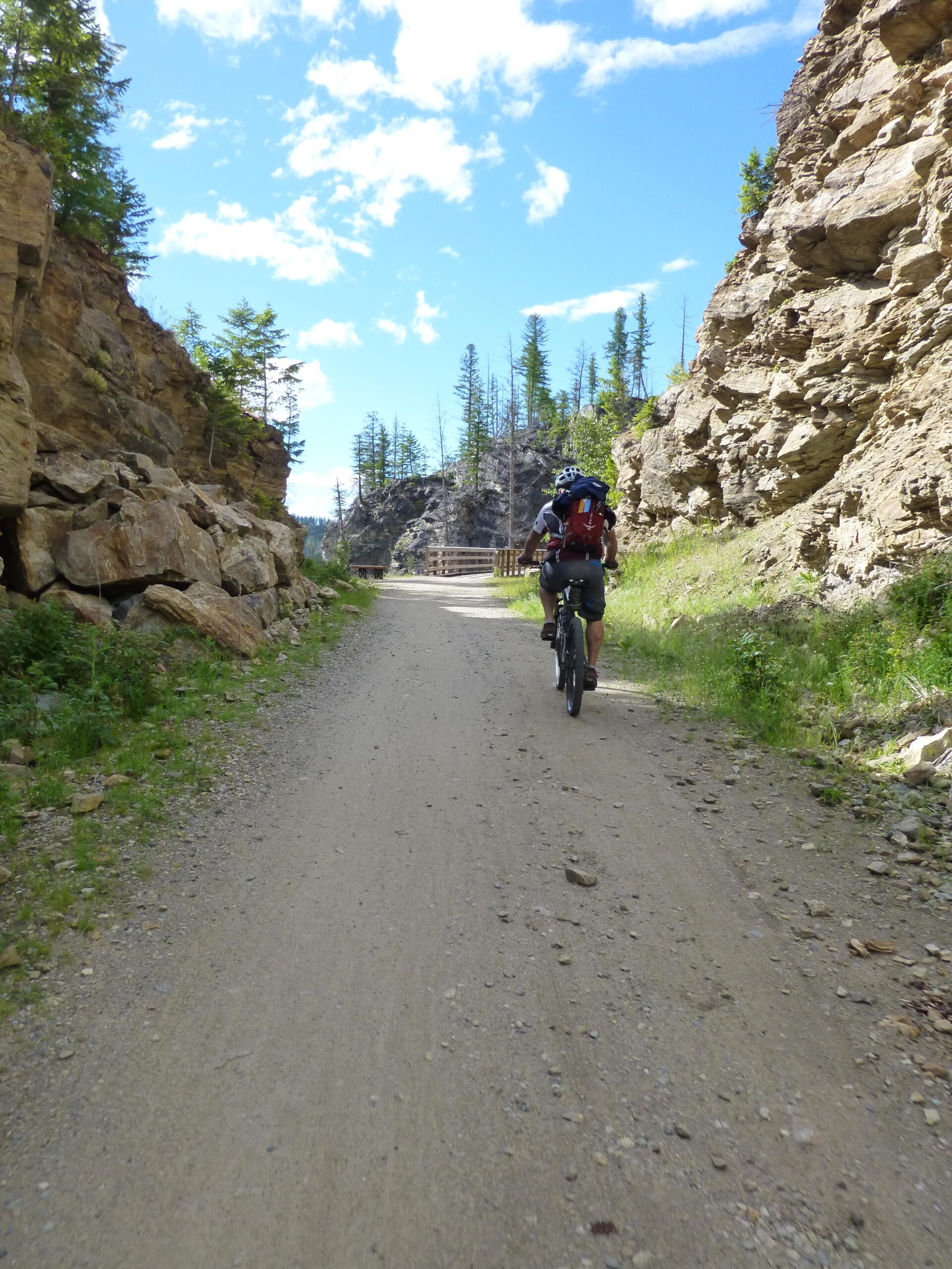A person riding a bicycle on a gravel trail surrounded by rocky cliffs and trees under a bright blue sky with scattered clouds. Kettle Valley Railway Trail (KVR) Myra Canyon to Penticton return mountain bike trail.
