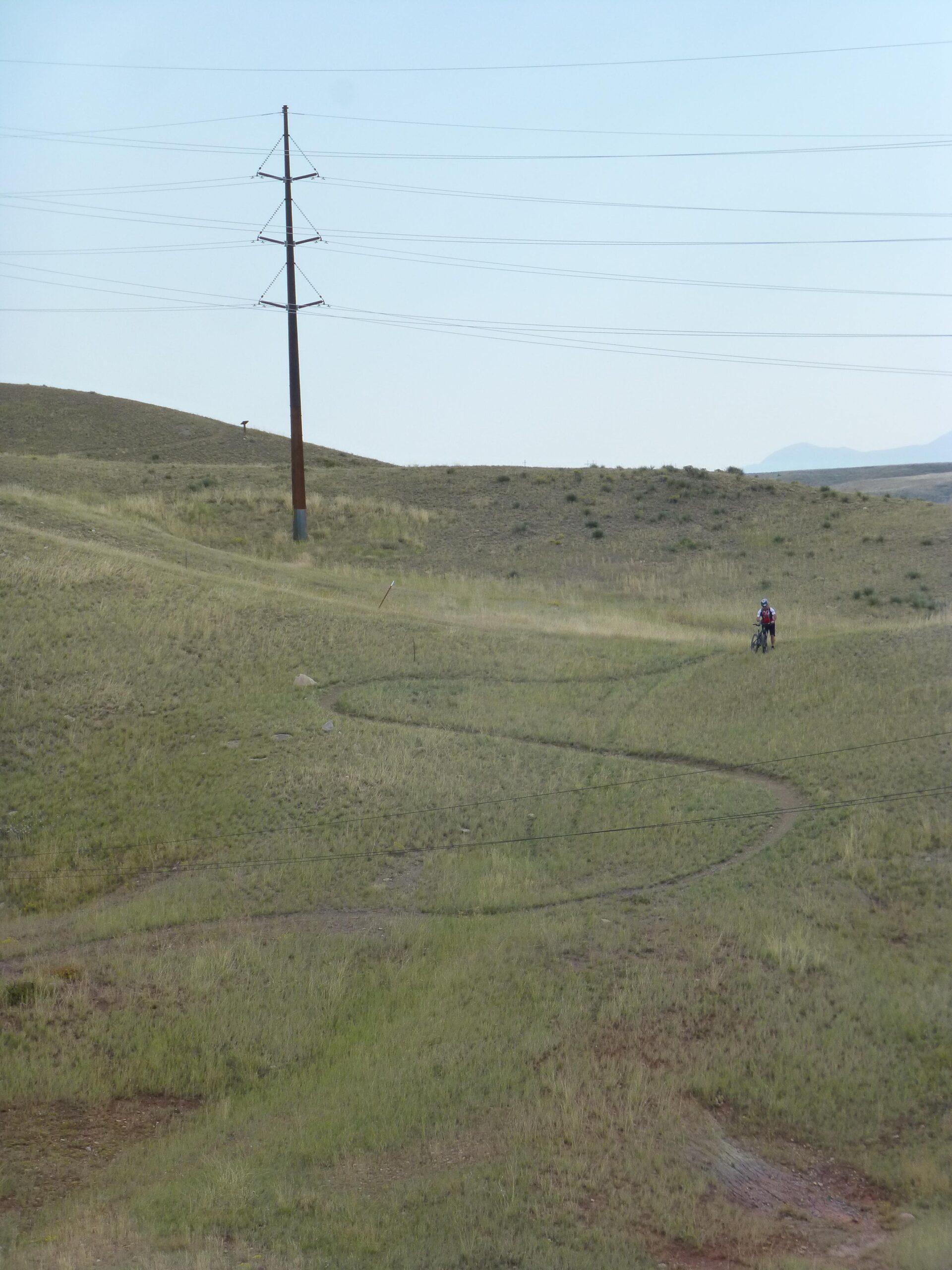 A winding dirt path through green hills under a clear sky, with a power line in the foreground and a cyclist riding in the distance. New Video Game/Yeah Kid mountain bike trail.