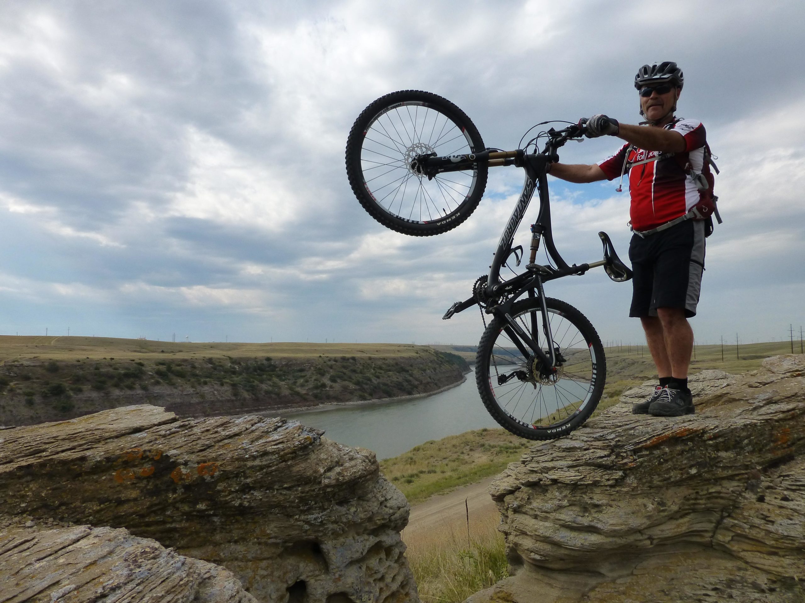 A cyclist in a red and black jersey stands on a rocky ledge, triumphantly lifting their mountain bike above their head. The background features a river and rolling hills under a cloudy sky, suggesting an adventurous outdoor setting. New Video Game/Yeah Kid mountain bike trail.