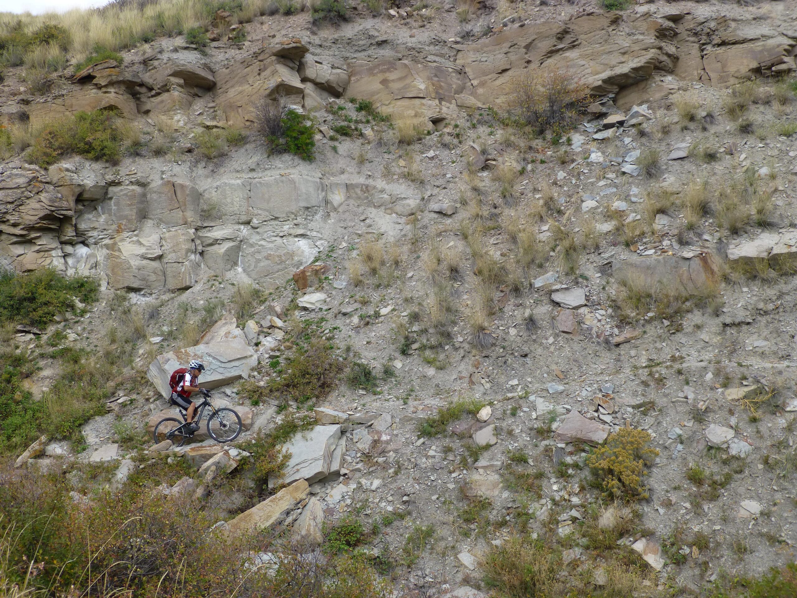 A mountain biker navigates a rocky hillside, surrounded by grass and shrubs. The terrain features loose stones and uneven ground, showcasing the challenges of off-road cycling. The background consists of layered rock formations, typical of a rugged landscape. New Video Game/Yeah Kid mountain bike trail.