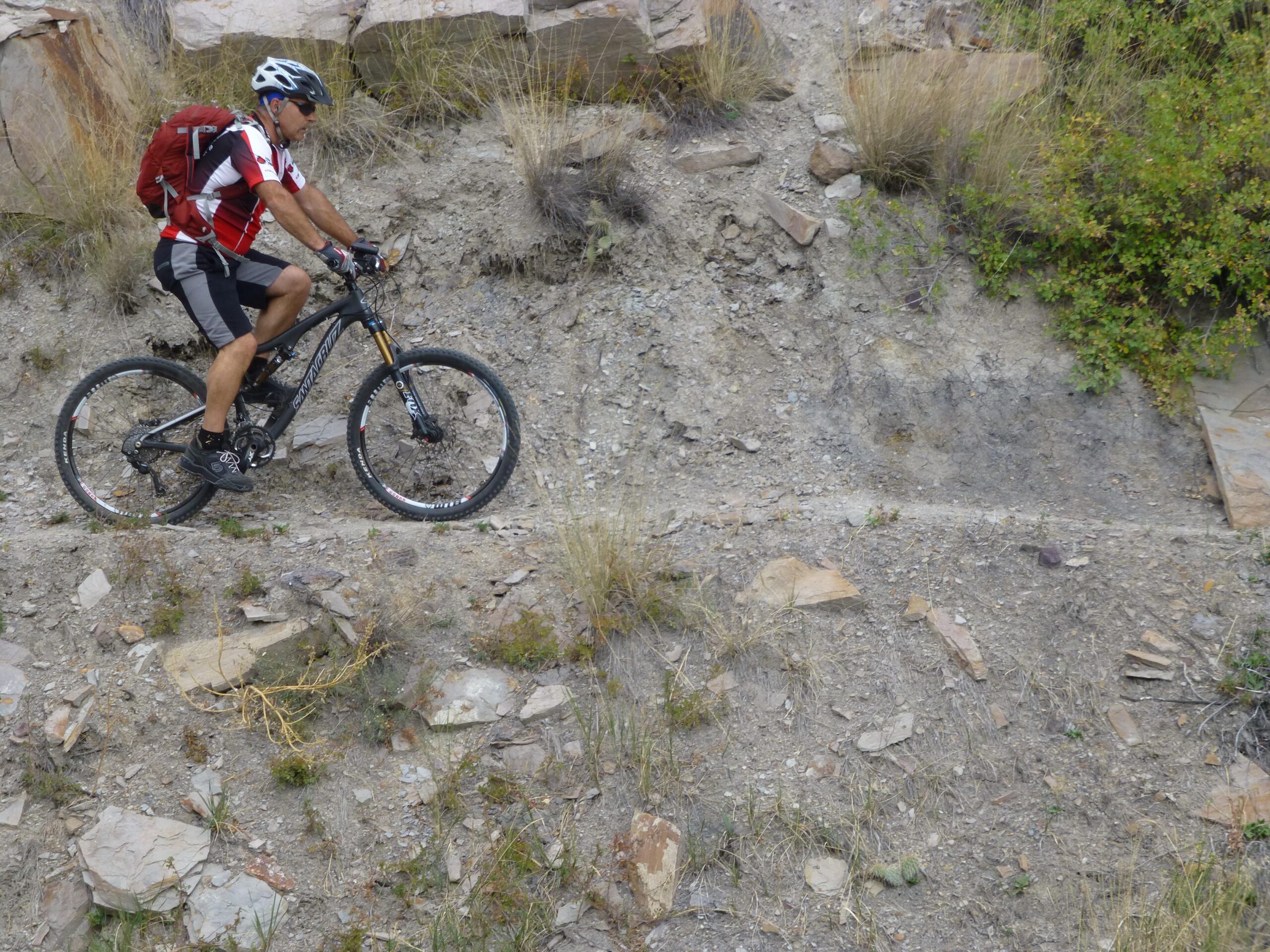 A mountain biker rides along a rocky, narrow trail in a rugged outdoor setting, wearing a helmet and a red and white biking jersey. He has a red backpack and is focused on navigating the uneven terrain surrounded by sparse vegetation and rocky outcrops. New Video Game/Yeah Kid mountain bike trail.