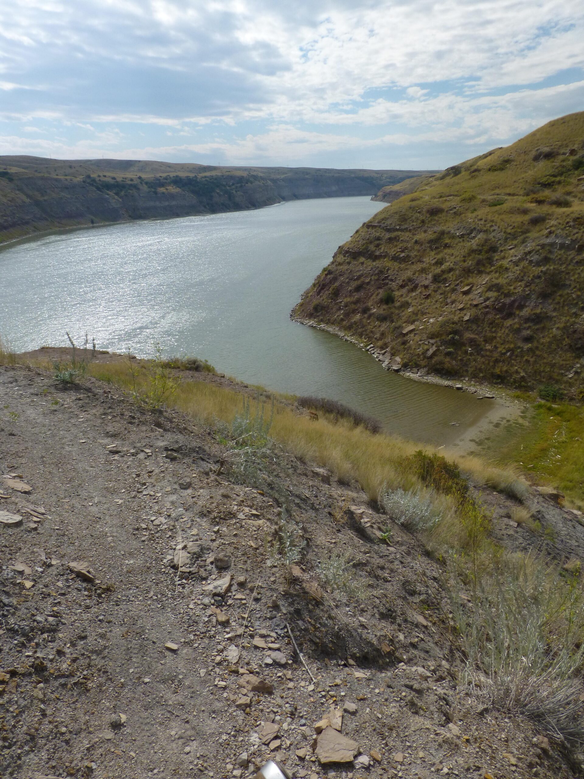 A scenic view of a winding river surrounded by grassy hills under a partly cloudy sky. The landscape features rocky terrain and sparse vegetation along the riverbank, with gentle slopes leading down to the water. New Video Game/Yeah Kid mountain bike trail.