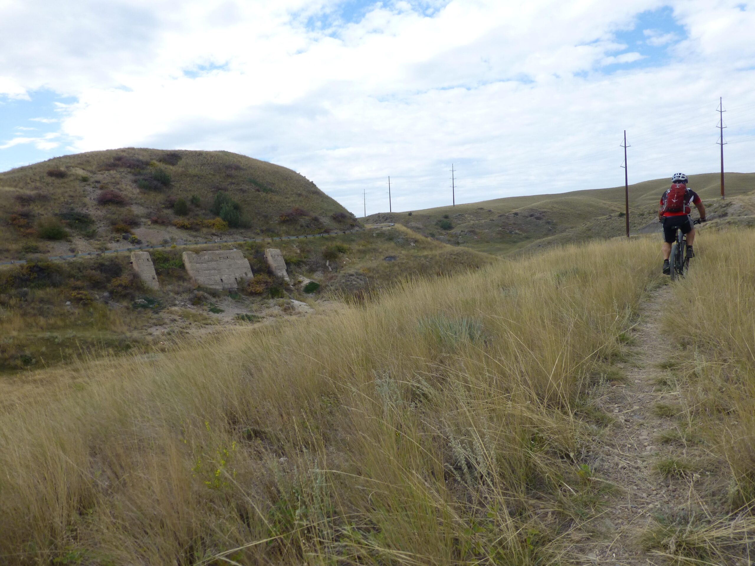 A cyclist rides along a narrow dirt path in a grassy landscape, with rolling hills and a cloudy sky in the background. Stone structures and power lines can be seen on the hillside, adding to the natural scenery. New Video Game/Yeah Kid mountain bike trail.