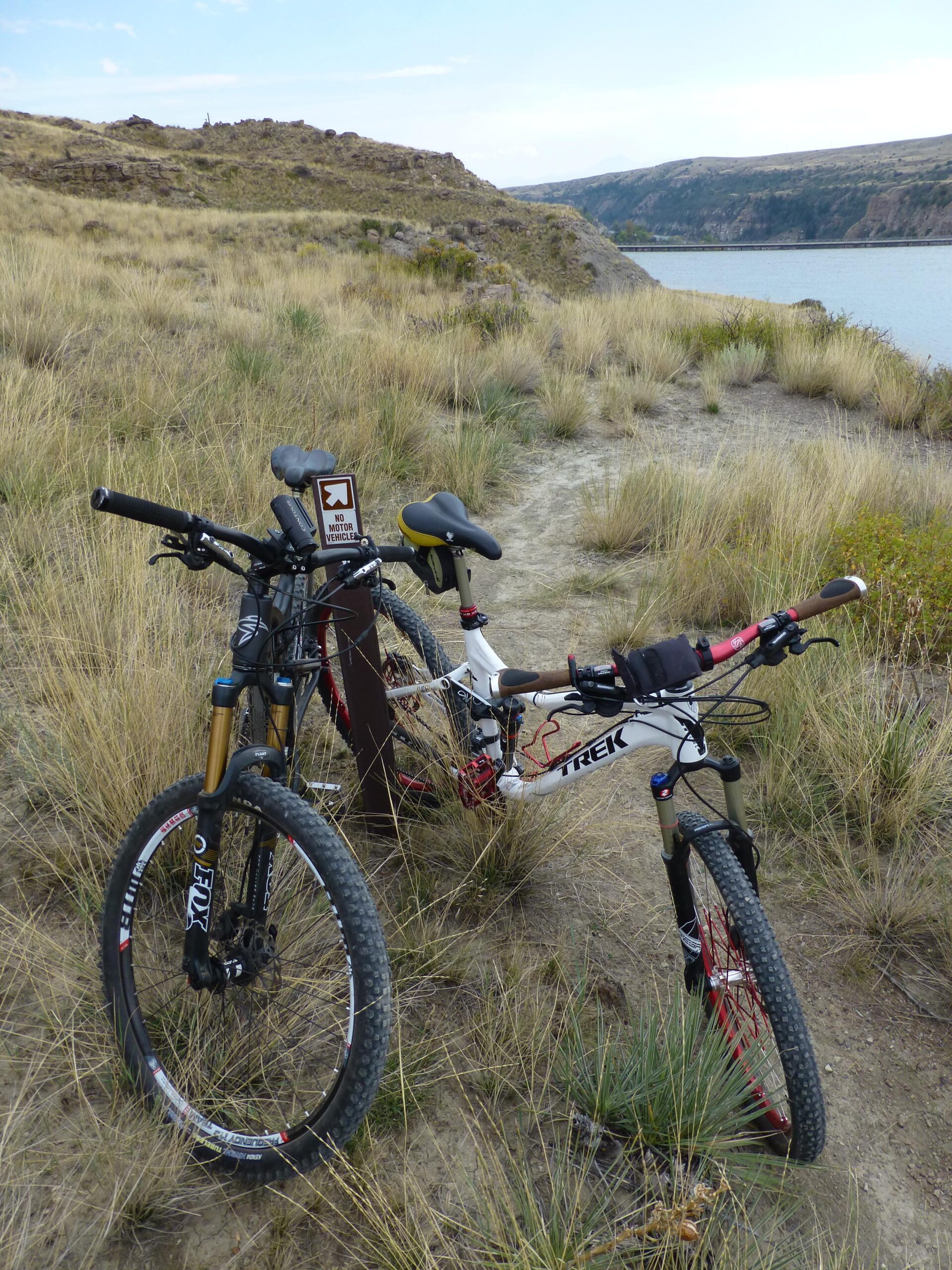 Two mountain bikes are parked on a grassy trail near a body of water, with rocky terrain and hills in the background. A sign indicating "No Motor Vehicles" is visible alongside the bikes, emphasizing the natural setting. The sky is partly cloudy, suggesting a pleasant outdoor environment. New Video Game/Yeah Kid mountain bike trail.
