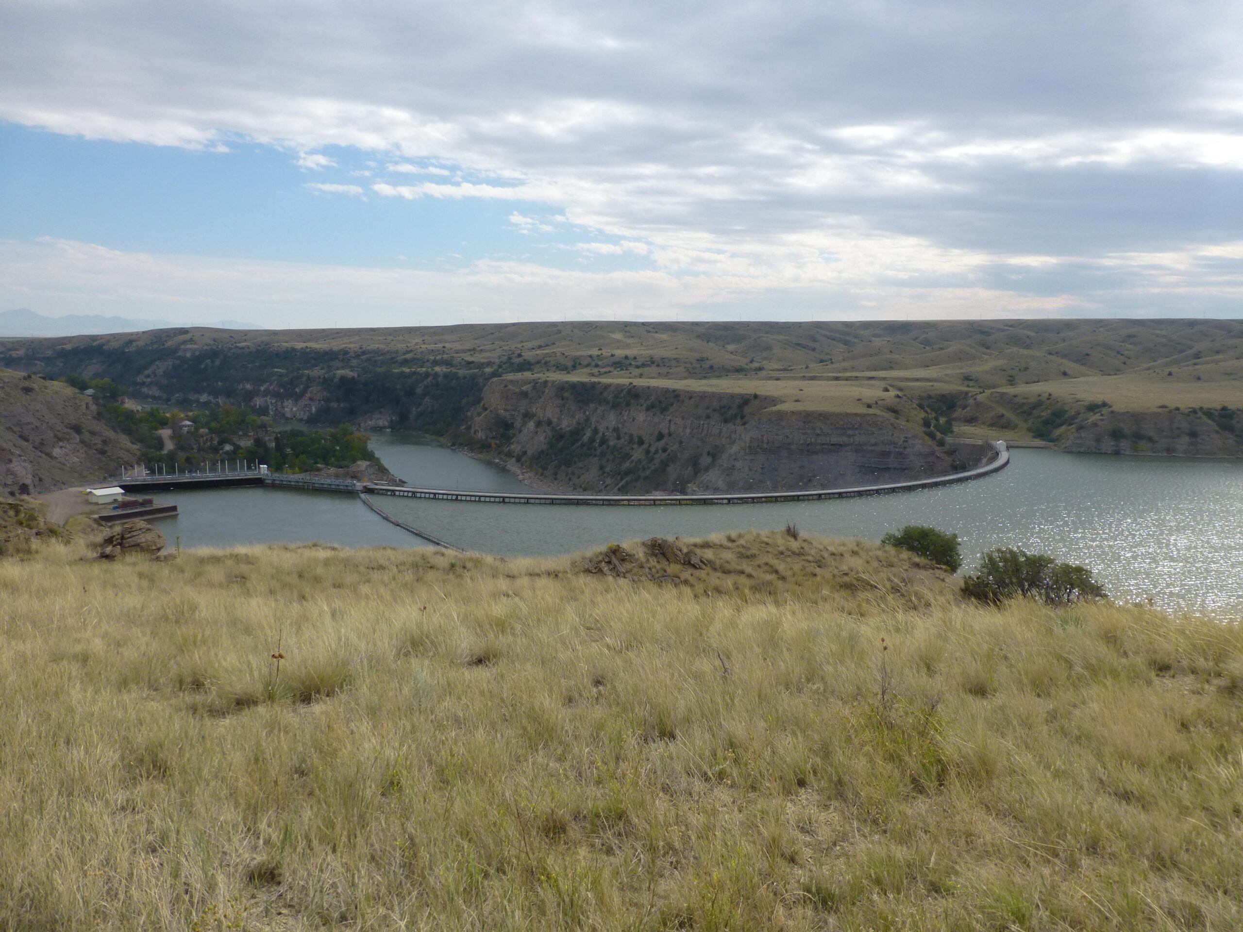 A scenic view of a reservoir nestled between rocky hills, with a winding dam and a pathway stretching across the water. The landscape features grassy slopes in the foreground and distant hills under a partly cloudy sky. New Video Game/Yeah Kid mountain bike trail.