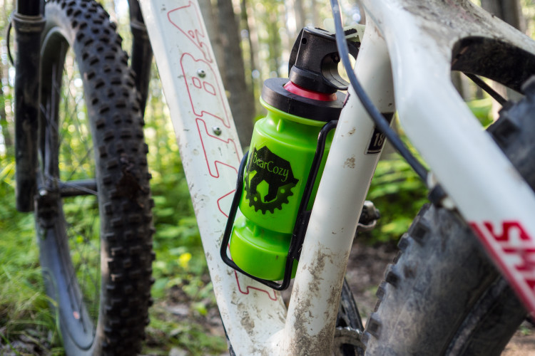 A green water bottle with a black holder is attached to the frame of a mountain bike. The background features a wooded area with trees and foliage, indicating an outdoor setting. The bike has thick tires suitable for off-road terrain.