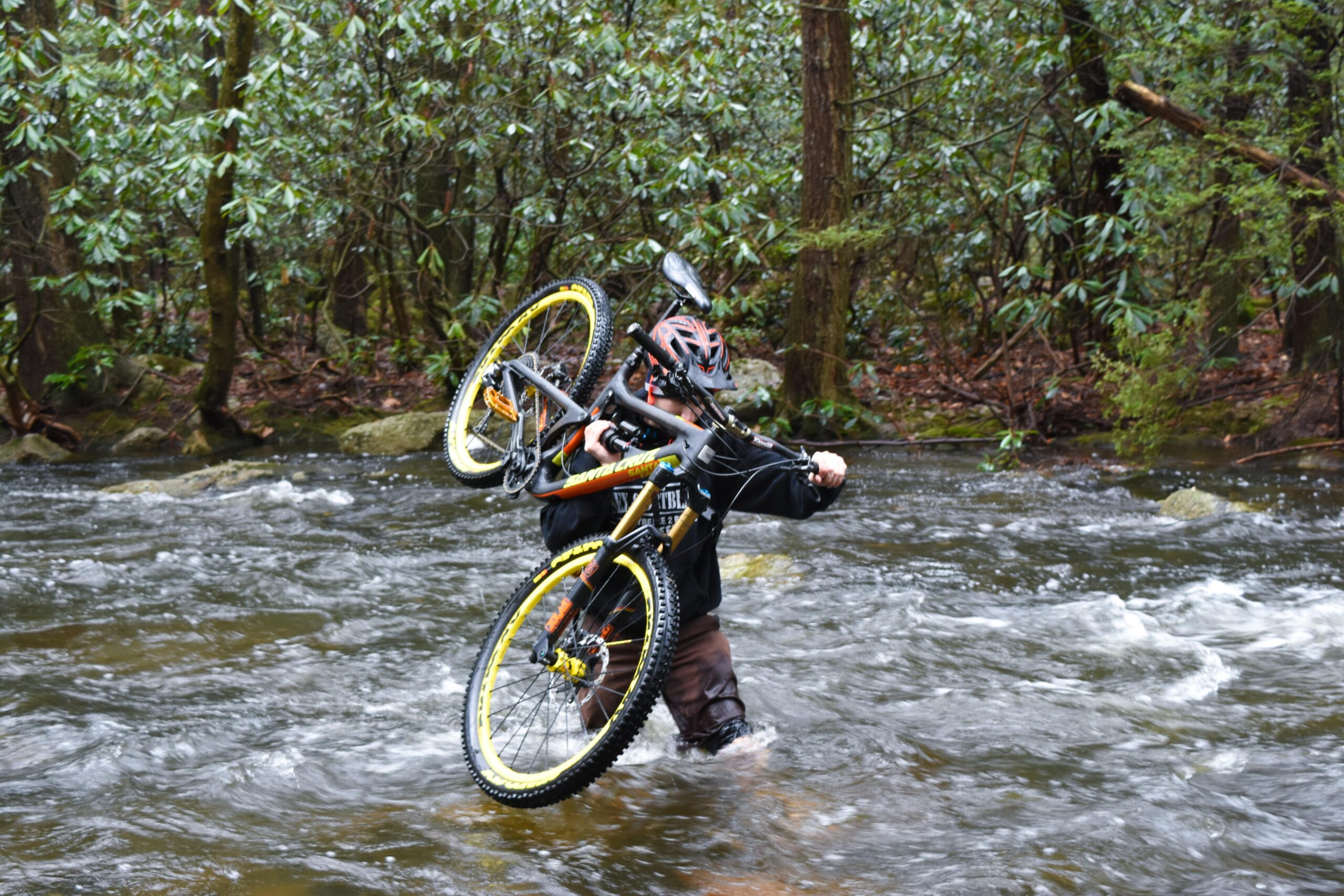 Santa Cruz Nomad Carbon 27.5: A person wearing a helmet and dark clothing is wading through a flowing stream while carrying a mountain bike over their shoulder. The surrounding area is lush with greenery and trees, indicating a natural, outdoor environment.