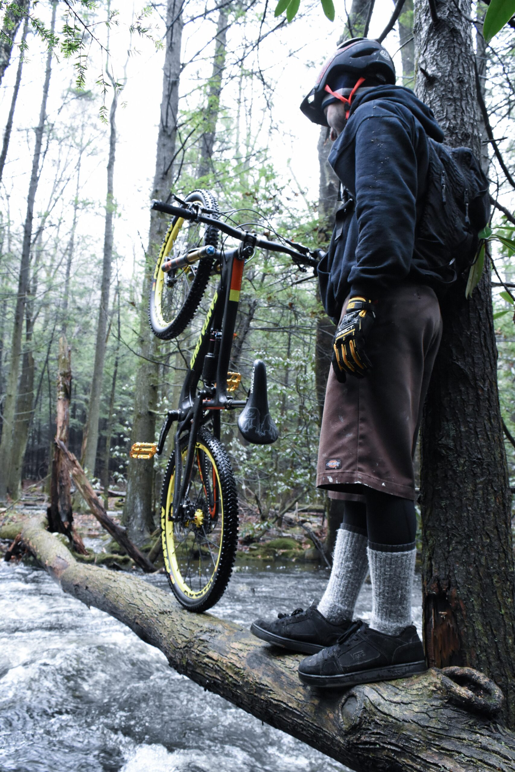 Santa Cruz Nomad Carbon 27.5: A mountain biker standing on a fallen tree over a stream in a forest, holding a bike upright. The rider is wearing a black helmet, a black hoodie, brown shorts, and long gray socks with black shoes. The background features dense trees and a flowing stream.