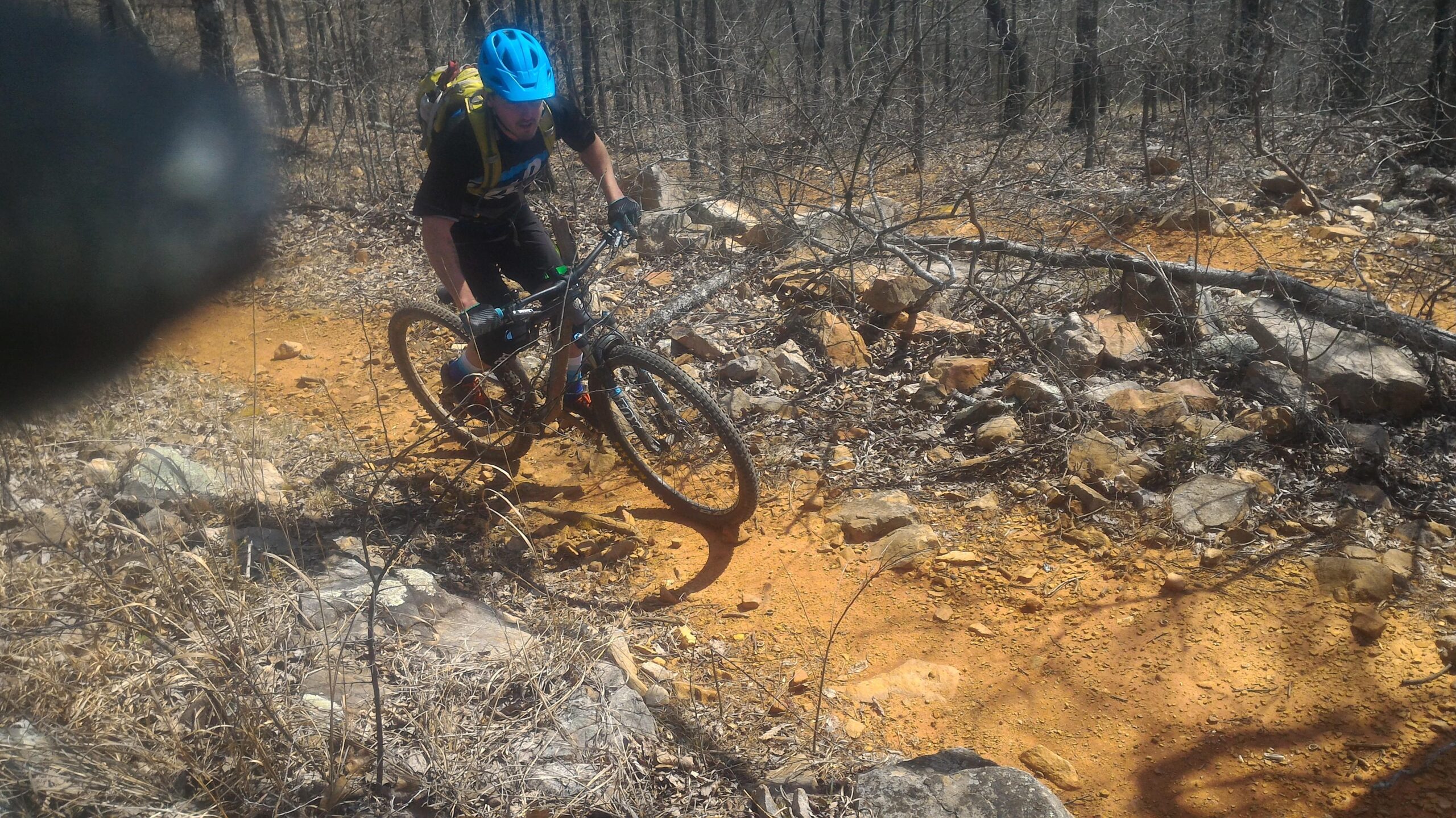 A mountain biker climbs a rocky, orange dirt trail, surrounded by bare trees and sparse vegetation. The rider wears a blue helmet and a backpack, focusing intently on navigating the trail. Coldwater Mountain mountain bike trail.