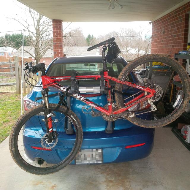 Giant Talon 27.5 4: A red mountain bike secured on the back of a blue car in a garage. The bike is muddy, indicating recent use, and the car is parked under a covered area with a brick wall nearby.
