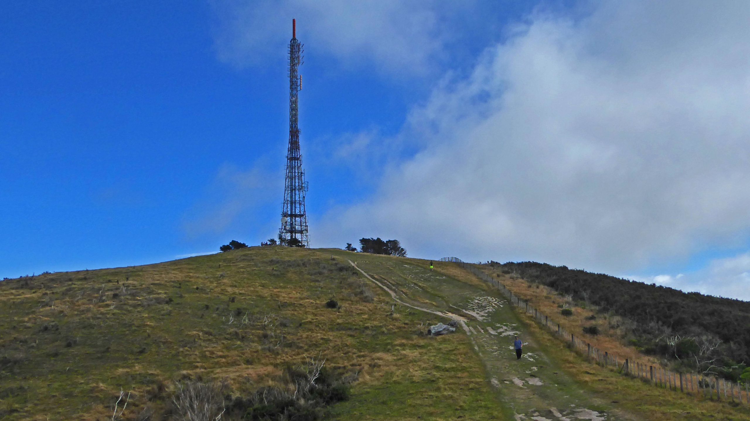 A tall communications tower stands on a grassy hill, with a clear blue sky and scattered clouds in the background. A person walks along a dirt path leading up the slope. Fencing borders the trail and the hillside, which features patches of shrubs and grass. Skyline Makara Loop mountain bike trail.