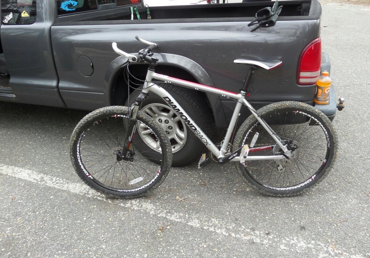 Diamondback Axis 27.5: A silver and black Diamondback mountain bike parked beside a gray pickup truck in a gravel parking lot. The bike features knobby tires and a minimalist design, with a water bottle in the truck's bed visible in the background. The scene suggests a recreational outdoor setting.