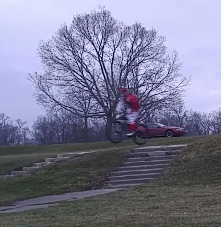 Specialized Hardrock Sport Disc: A person dressed in red clothing riding a bicycle, mid-jump over a set of steps in a park, with a bare tree and a red car in the background under a cloudy sky.