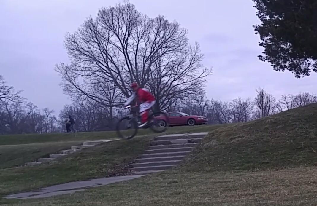 Specialized Hardrock Sport Disc: A person dressed in a red outfit rides a mountain bike down a grassy hill, catching air as they jump off a set of concrete steps. In the background, a red car is parked near a tree, with a person walking on the path. The sky is overcast, suggesting a cloudy day.