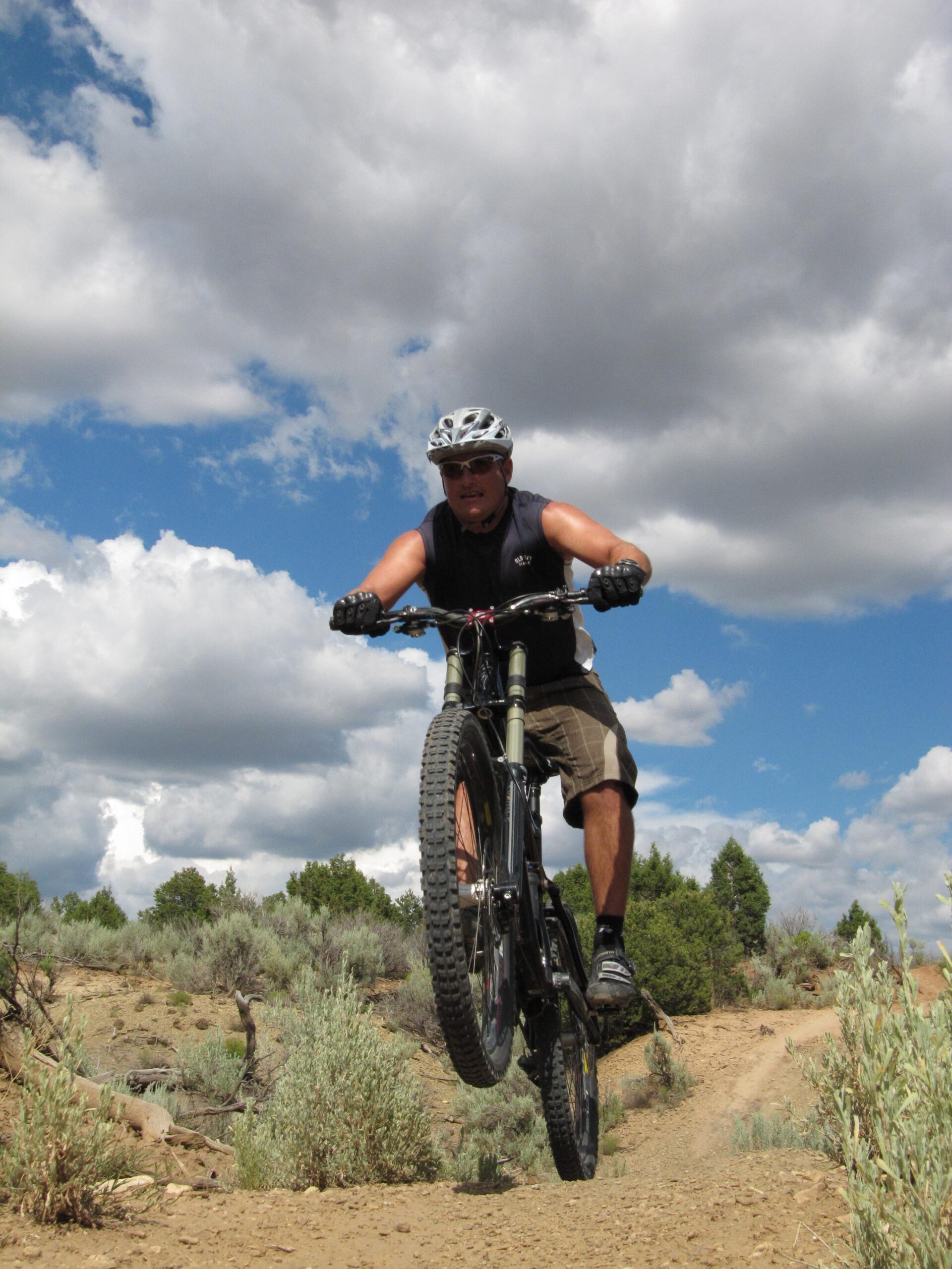 Specialized S-Works Enduro SL Carbon: A person riding a mountain bike on a dirt trail, performing a jump. They are wearing a helmet and sunglasses, with a focused expression. The background features a blue sky with fluffy clouds and vegetation typical of a desert or semi-arid environment.