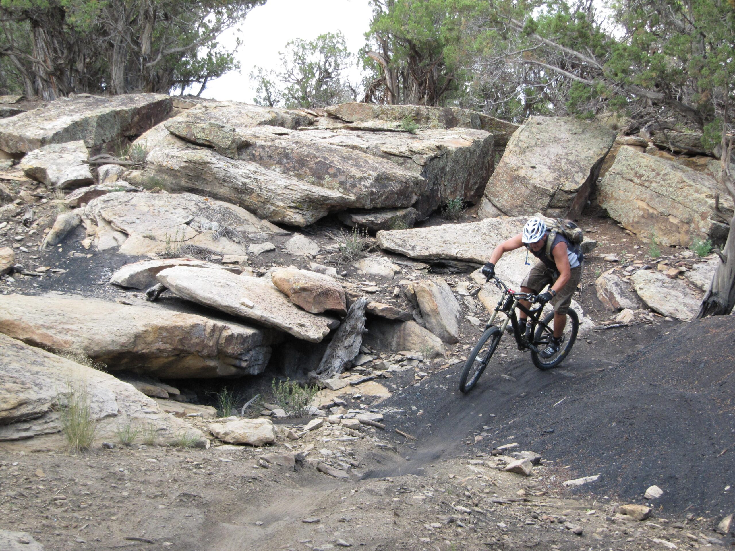 Specialized S-Works Enduro SL Carbon: A mountain biker navigates rocky terrain on a trail, surrounded by large boulders and sparse vegetation. The rider is wearing a helmet and backpack, showcasing a dynamic posture as they ride downhill.
