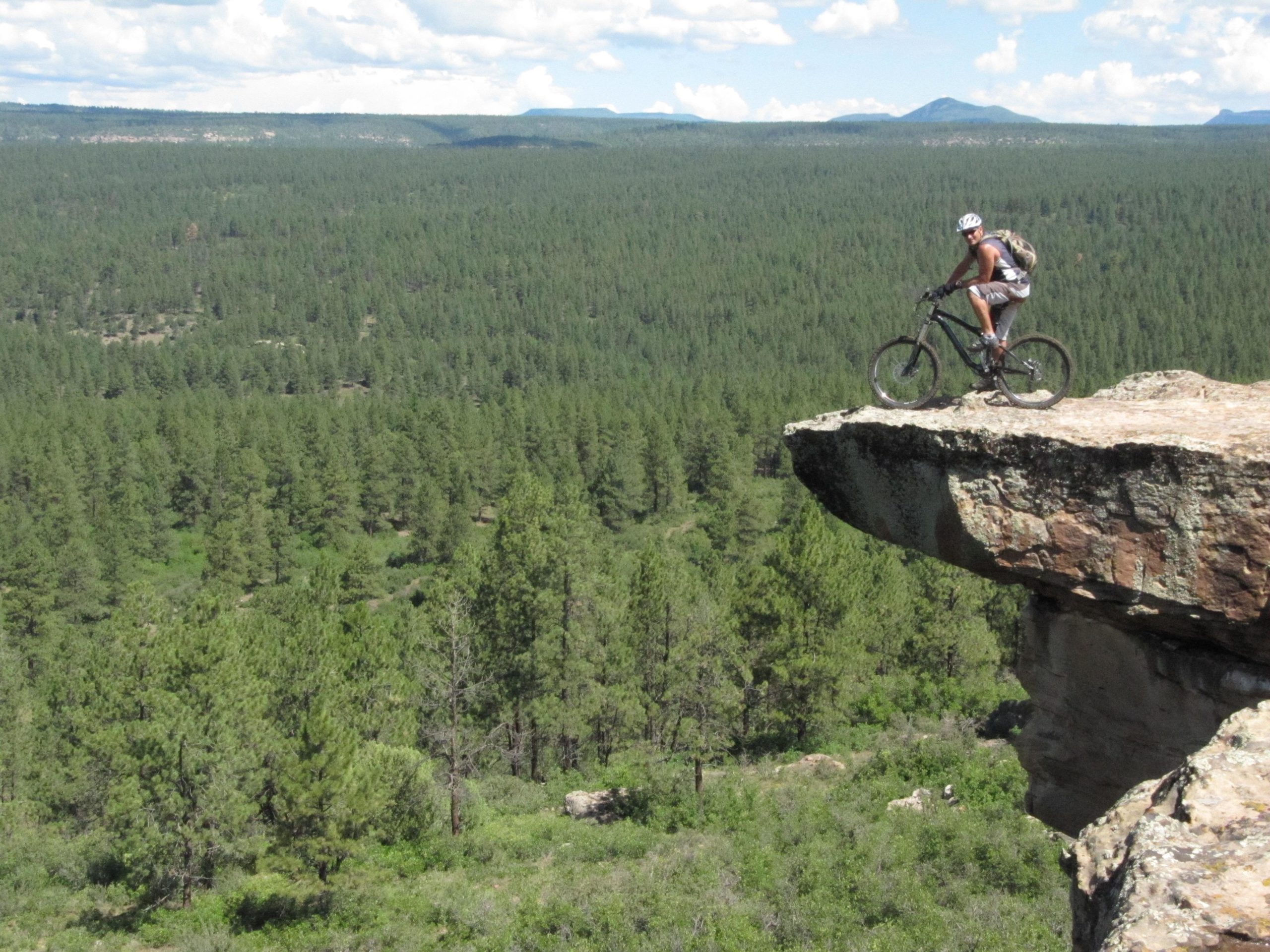 A mountain biker stands on the edge of a rocky cliff, overlooking a vast forest of tall green pine trees. The sunny sky is partly cloudy, with distant mountains visible on the horizon. Bean Canyon Loop mountain bike trail.