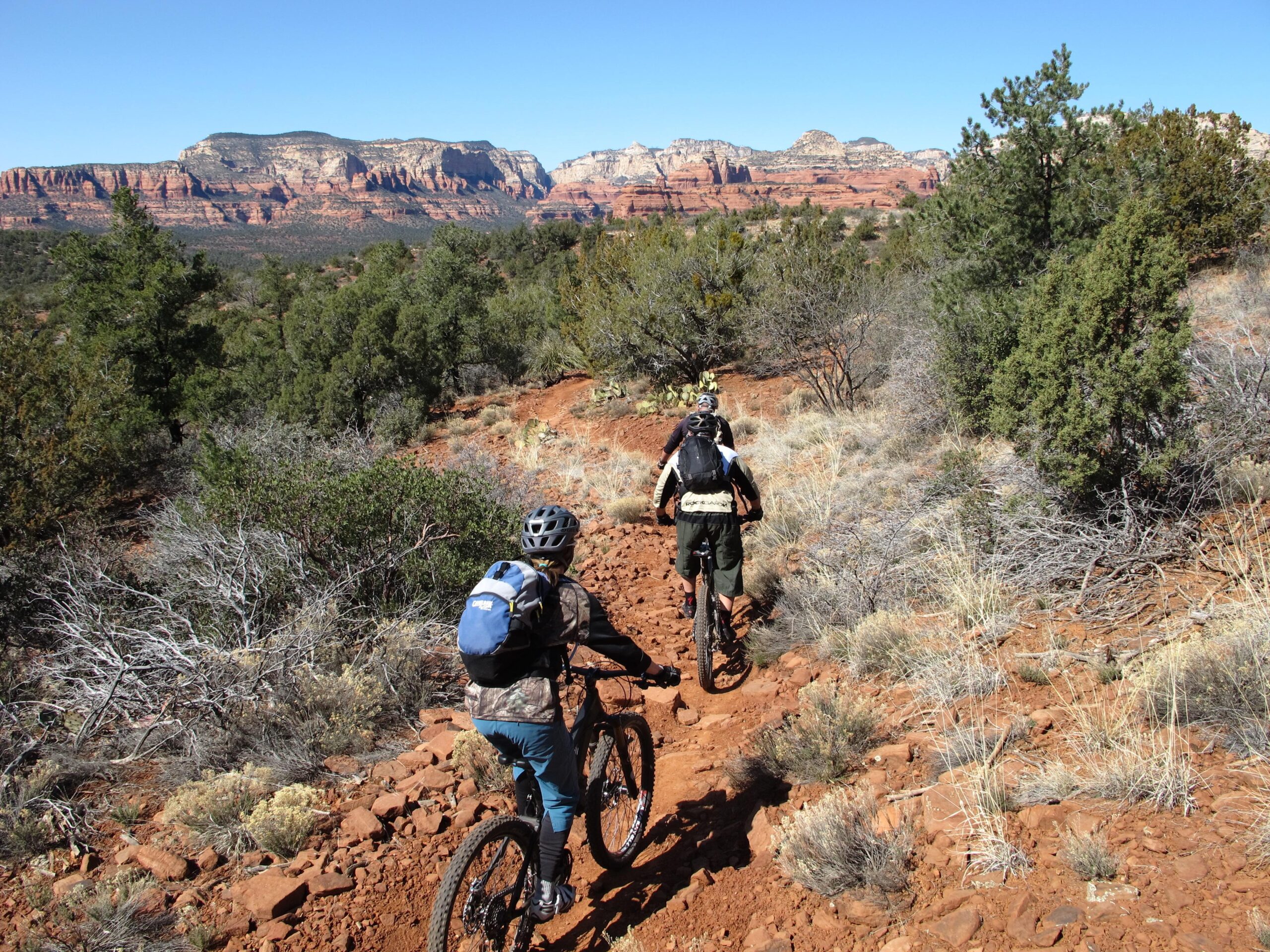 Two mountain bikers riding on a dirt trail in a rugged desert landscape, with red rock formations and sparse vegetation in the background under a clear blue sky. Bell Rock Area Trails mountain bike trail.