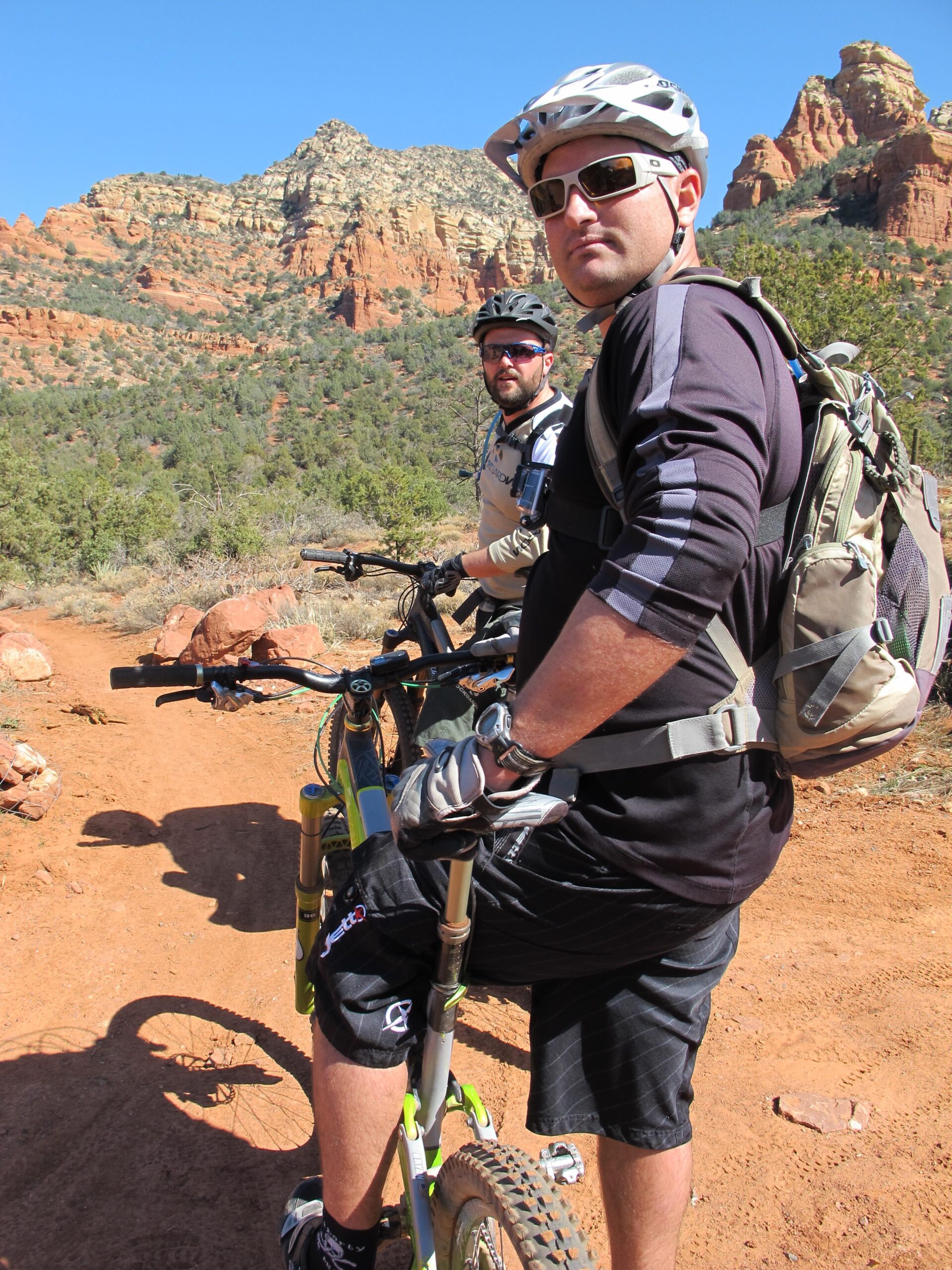 Two mountain bikers pause on a dirt trail surrounded by rocky terrain and greenery. The first biker, wearing a black shirt and a backpack, faces the camera, while the second biker, in a light-colored shirt and sunglasses, looks slightly off to the side. The background features striking red rock formations under a clear blue sky. Bell Rock Area Trails mountain bike trail.