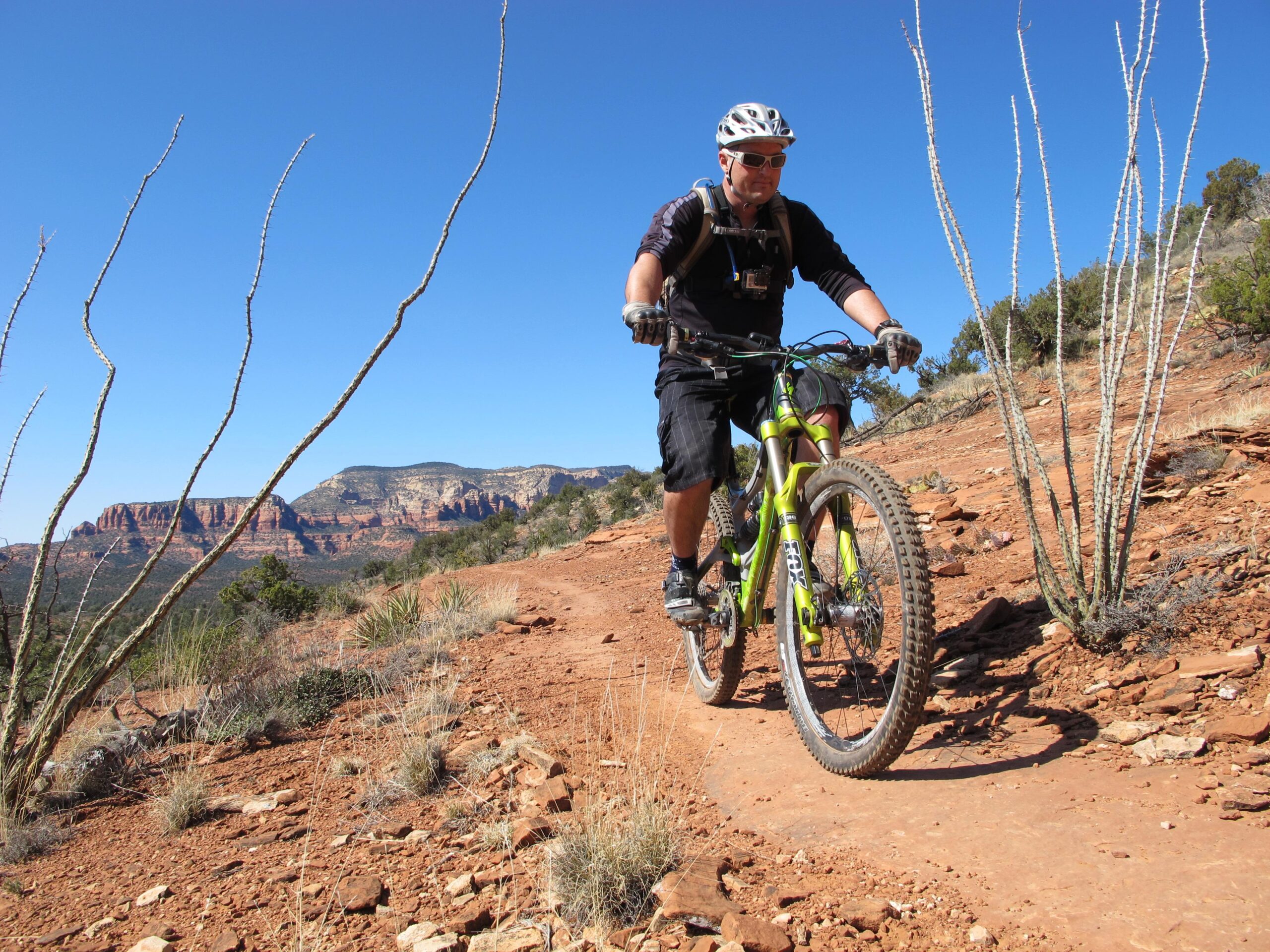 A mountain biker riding along a rocky trail in a desert landscape, with a backdrop of red rock formations under a clear blue sky. The biker is wearing a helmet and sunglasses, equipped with a backpack and riding a green mountain bike. Cacti and desert vegetation are visible on either side of the path. Bell Rock Area Trails mountain bike trail.