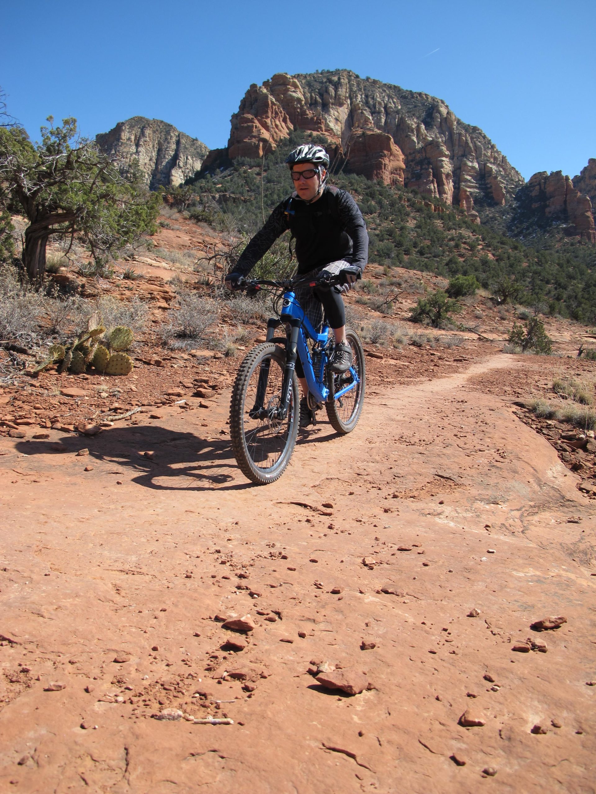 A cyclist in a black long-sleeve shirt and helmet rides a blue mountain bike along a rocky dirt trail. Red rock formations and sparse vegetation are visible in the background under a clear blue sky. Bell Rock Area Trails mountain bike trail.