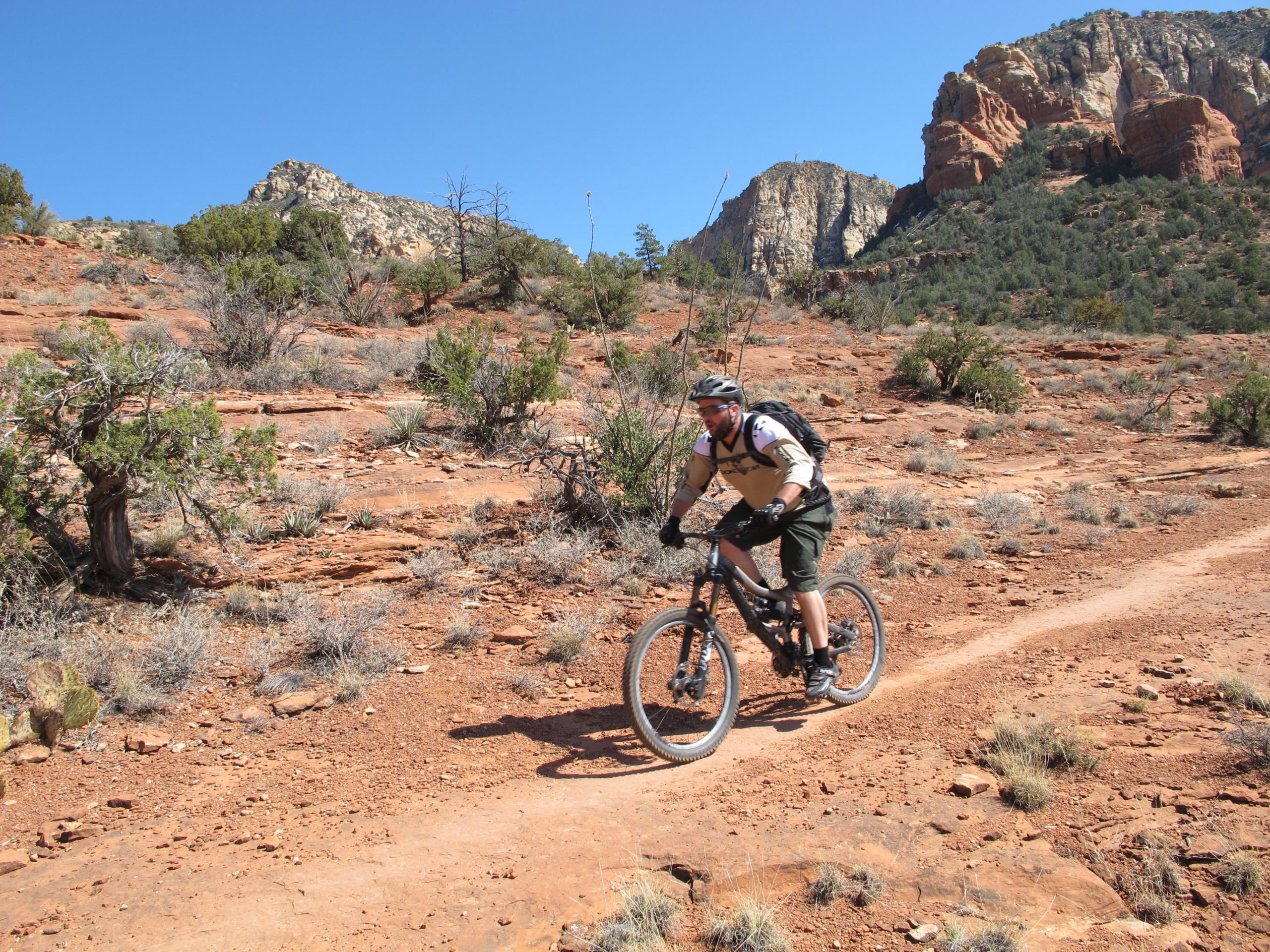A person riding a mountain bike on a rocky trail surrounded by desert vegetation, with red rock formations and blue sky in the background. Bell Rock Area Trails mountain bike trail.